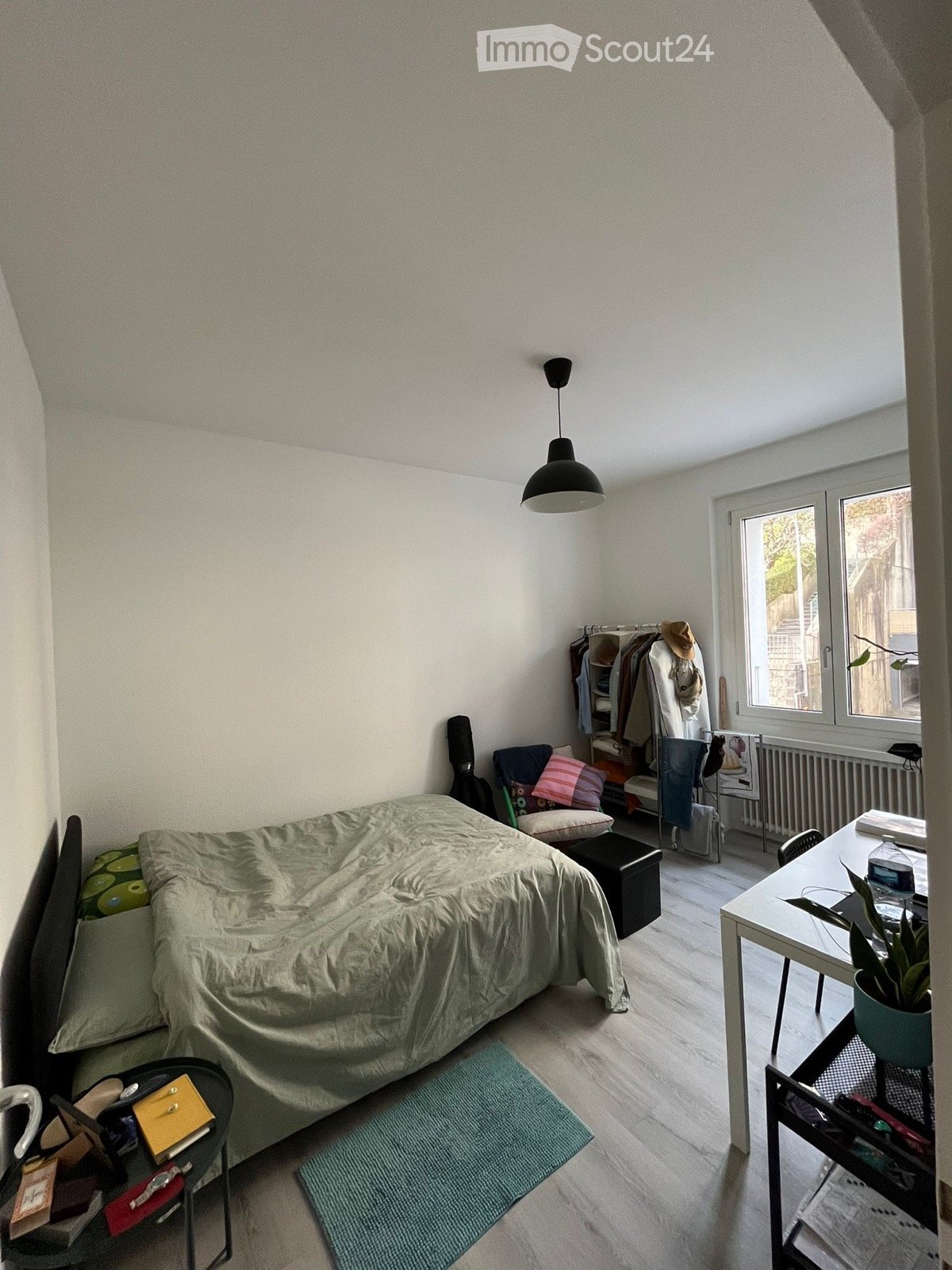 A small, well-lit bedroom with a bed, a desk, a chair, a stool, a plant, a radiator, a guitar, and a wardrobe.