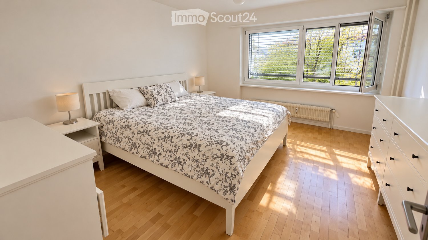 Bright bedroom with wooden flooring, large window with blinds, white bed with patterned bedding, nightstands with lamps, and a dresser.