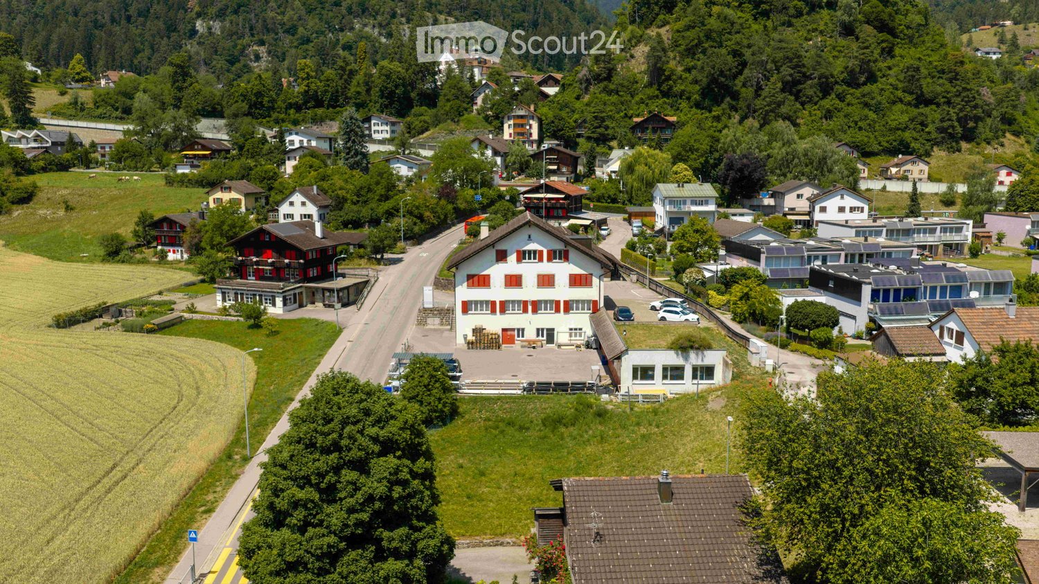 Aerial view of a rural village with multiple houses, surrounded by green fields and forested hills.