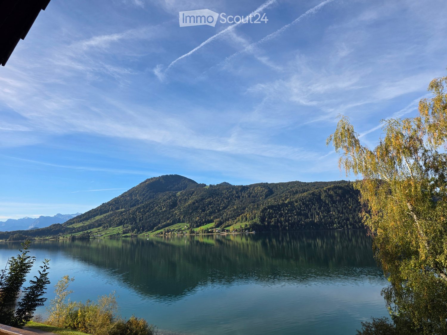 A serene lakeside view with calm water reflecting the surrounding mountains and trees, under a clear blue sky.
