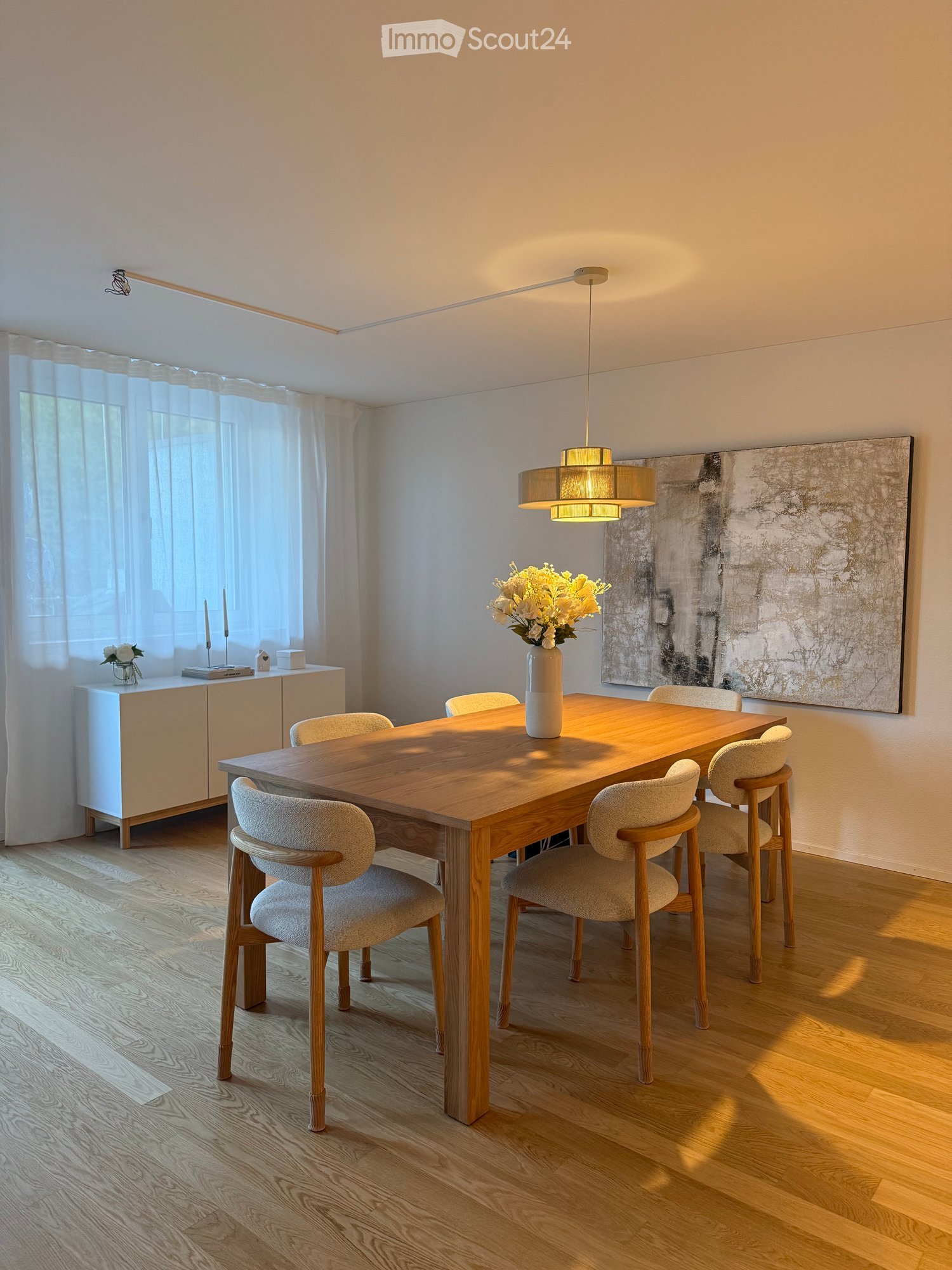 Modern dining room with a wooden dining table, six chairs, a vase with flowers, a cabinet, and a large abstract painting on the wall.