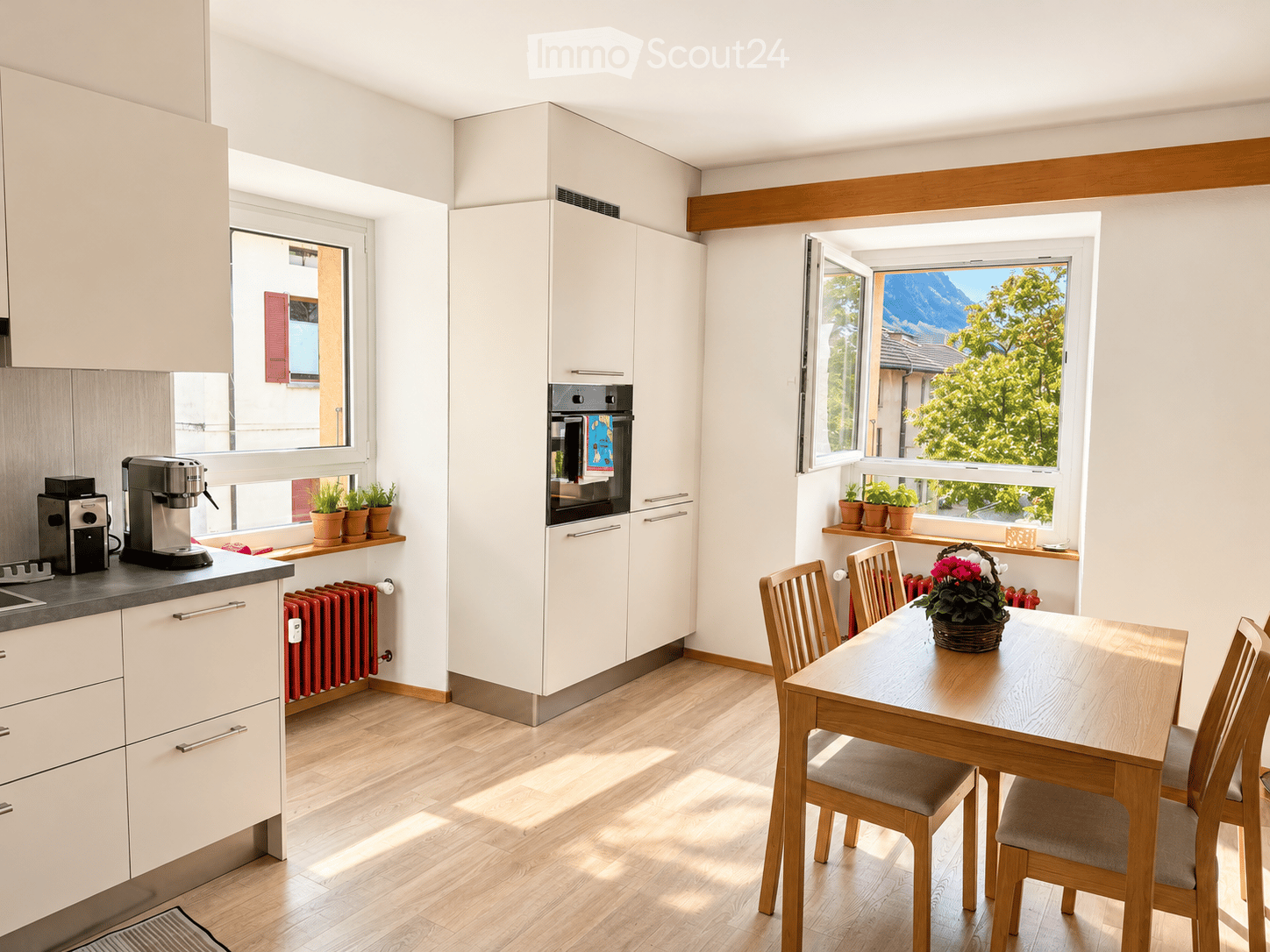 Bright kitchen with white cabinets, wooden floor, coffee maker, oven, dining table with chairs, and potted plants on windowsills.