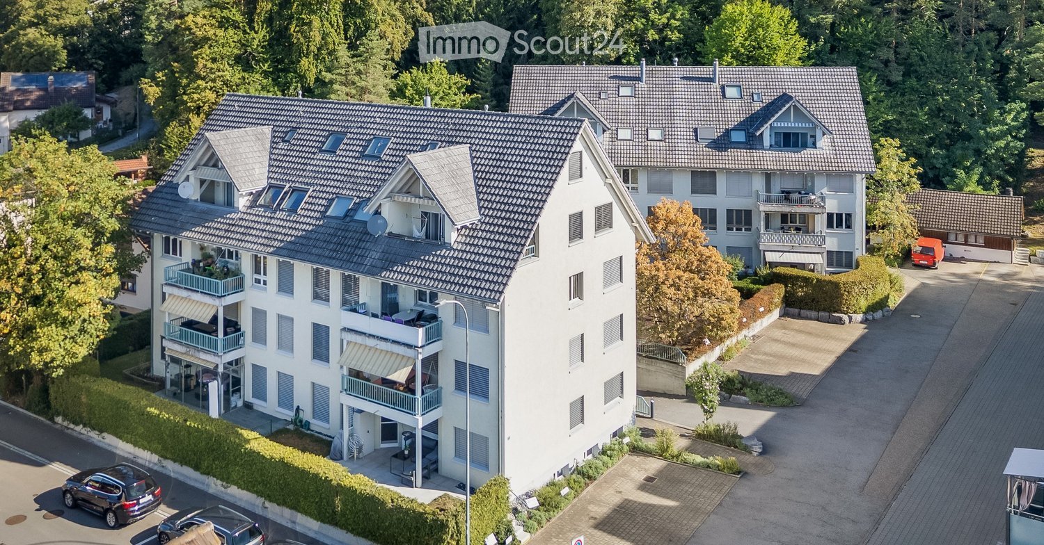 Aerial view of a multi-story residential building with multiple balconies, surrounded by greenery and parking spaces.