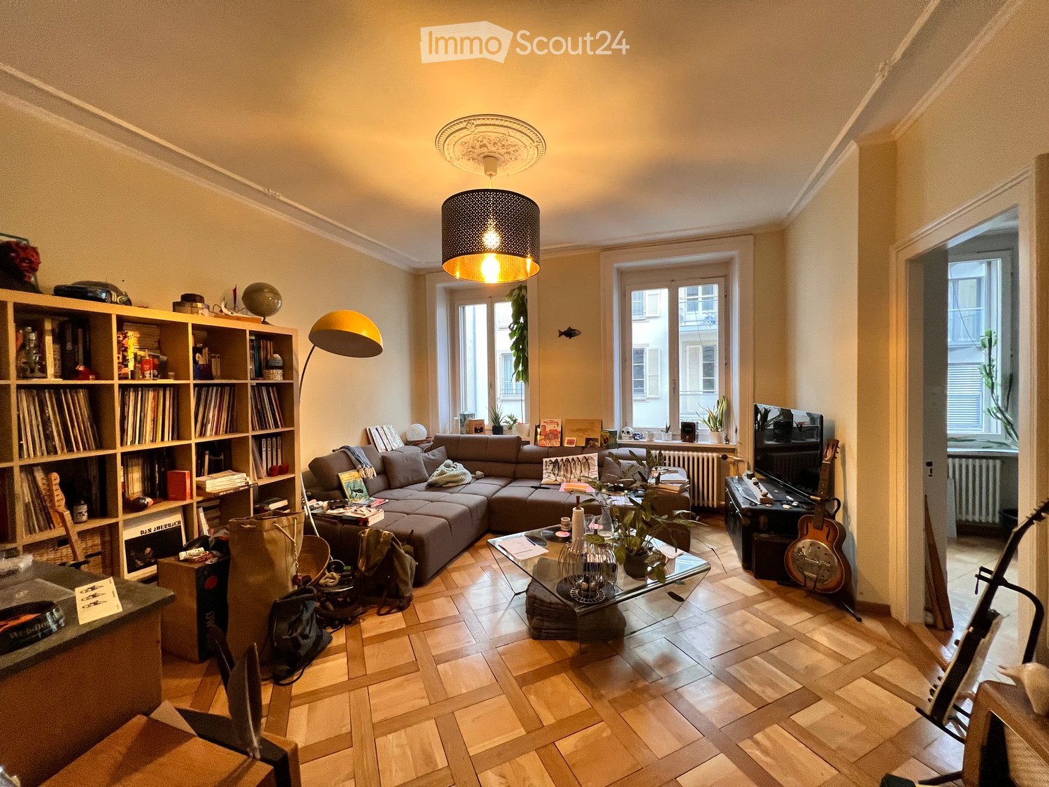 A cozy living room with a sectional sofa, coffee table, bookshelf, floor lamp, and guitar. The room features wooden flooring and large windows letting in natural light.