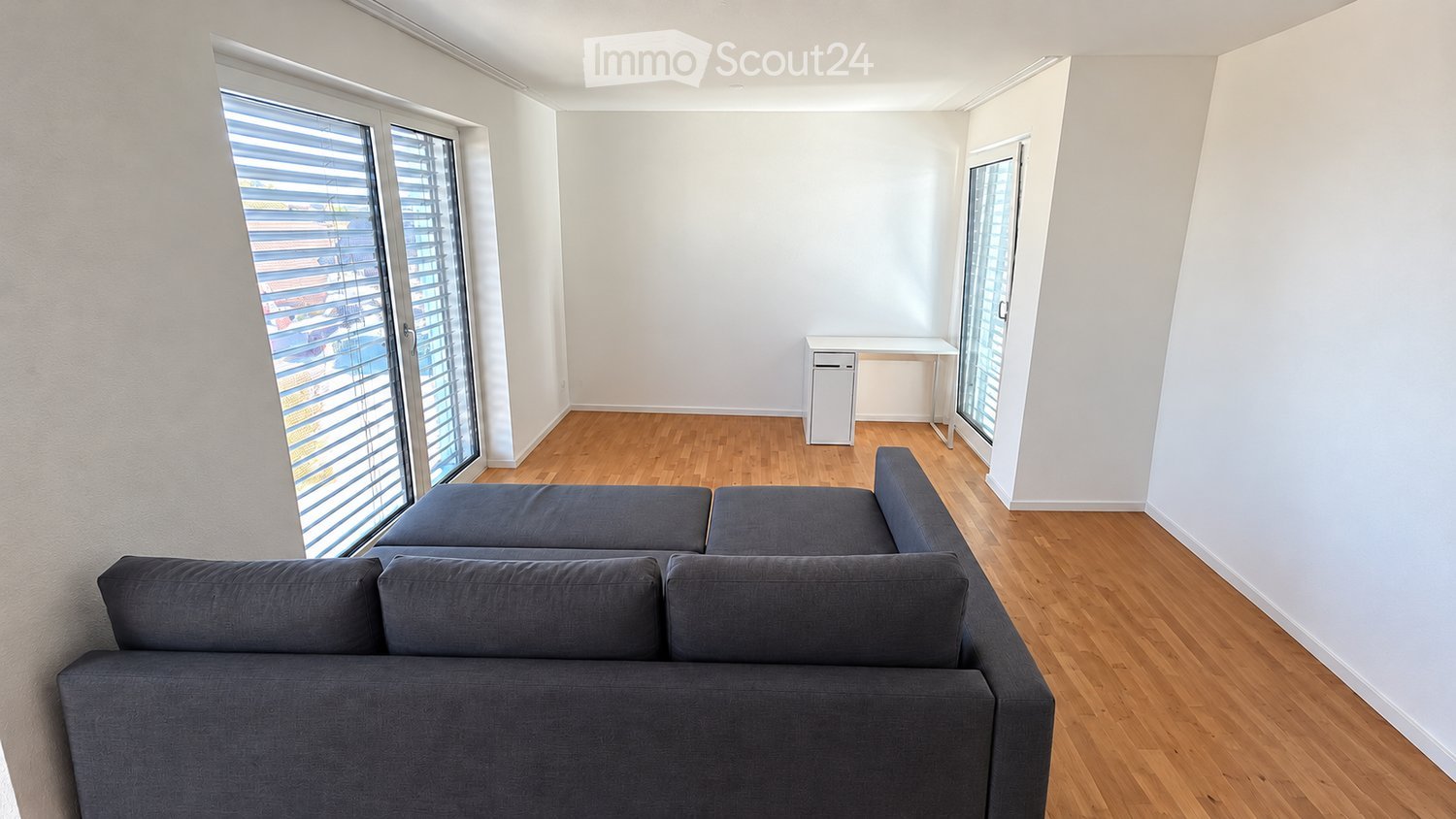 Bright living room with wooden flooring, large windows with blinds, grey sectional sofa, small desk, and chair.