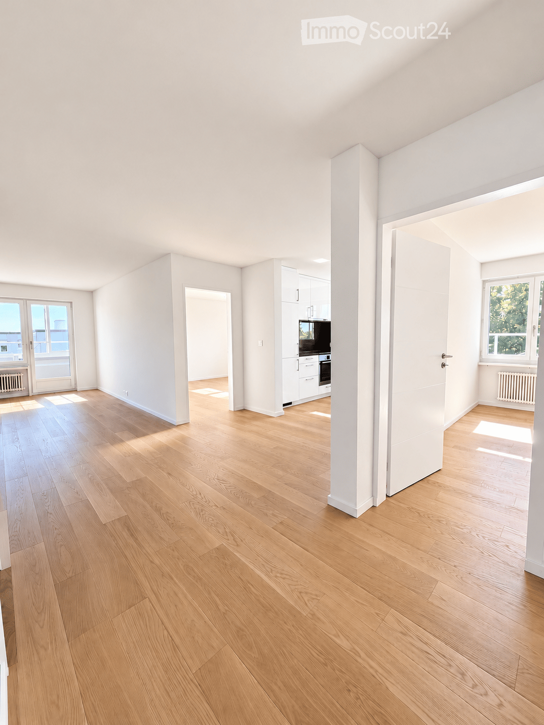 Bright, empty room with light wooden flooring, white walls, and large windows allowing natural light. The space features a central column and an open doorway leading to another area with white cabinetry and appliances.