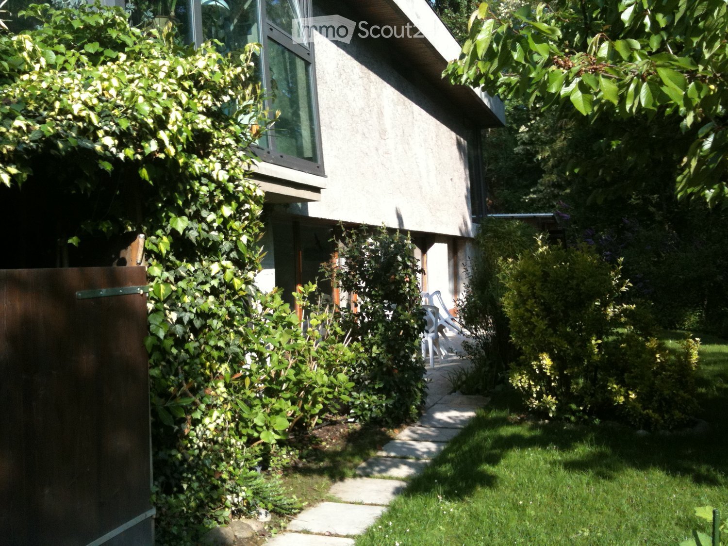 A modern house with a well-maintained garden, featuring a stone pathway, lush greenery, and a small seating area with white chairs near the entrance.