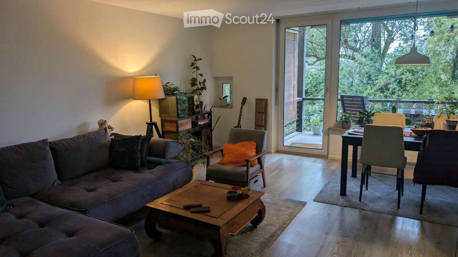 Bright living room with a grey sofa, wooden coffee table, armchair, dining table, and balcony access. Features include a guitar, floor lamp, and plants.