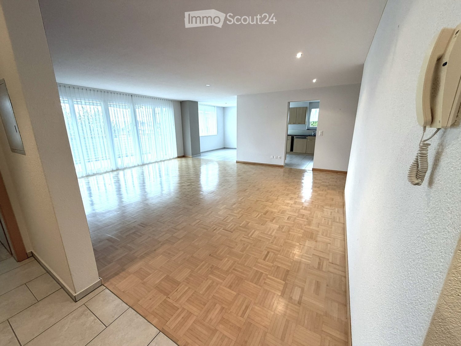 Empty room with wooden herringbone flooring, white walls, large windows with blinds, and a visible kitchen area in the background.