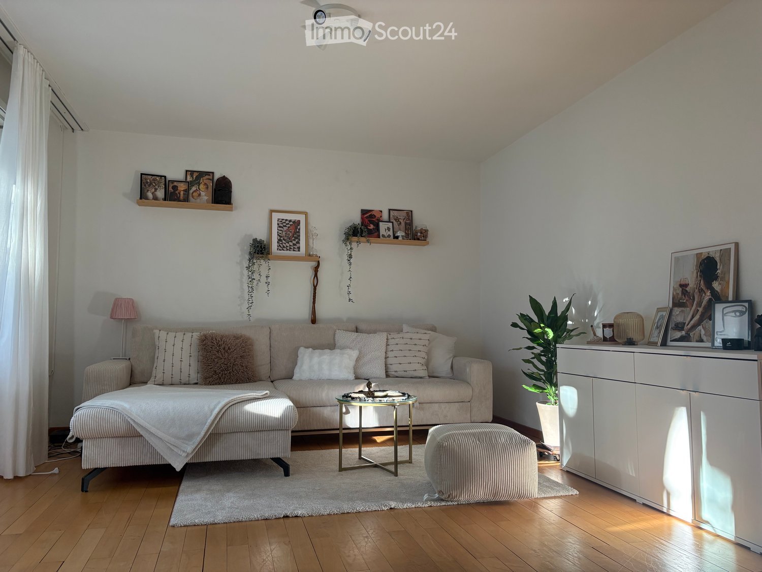 Modern living room with a beige sofa, coffee table, ottoman, side table, plant, cabinet, and shelves with picture frames and decorative items.