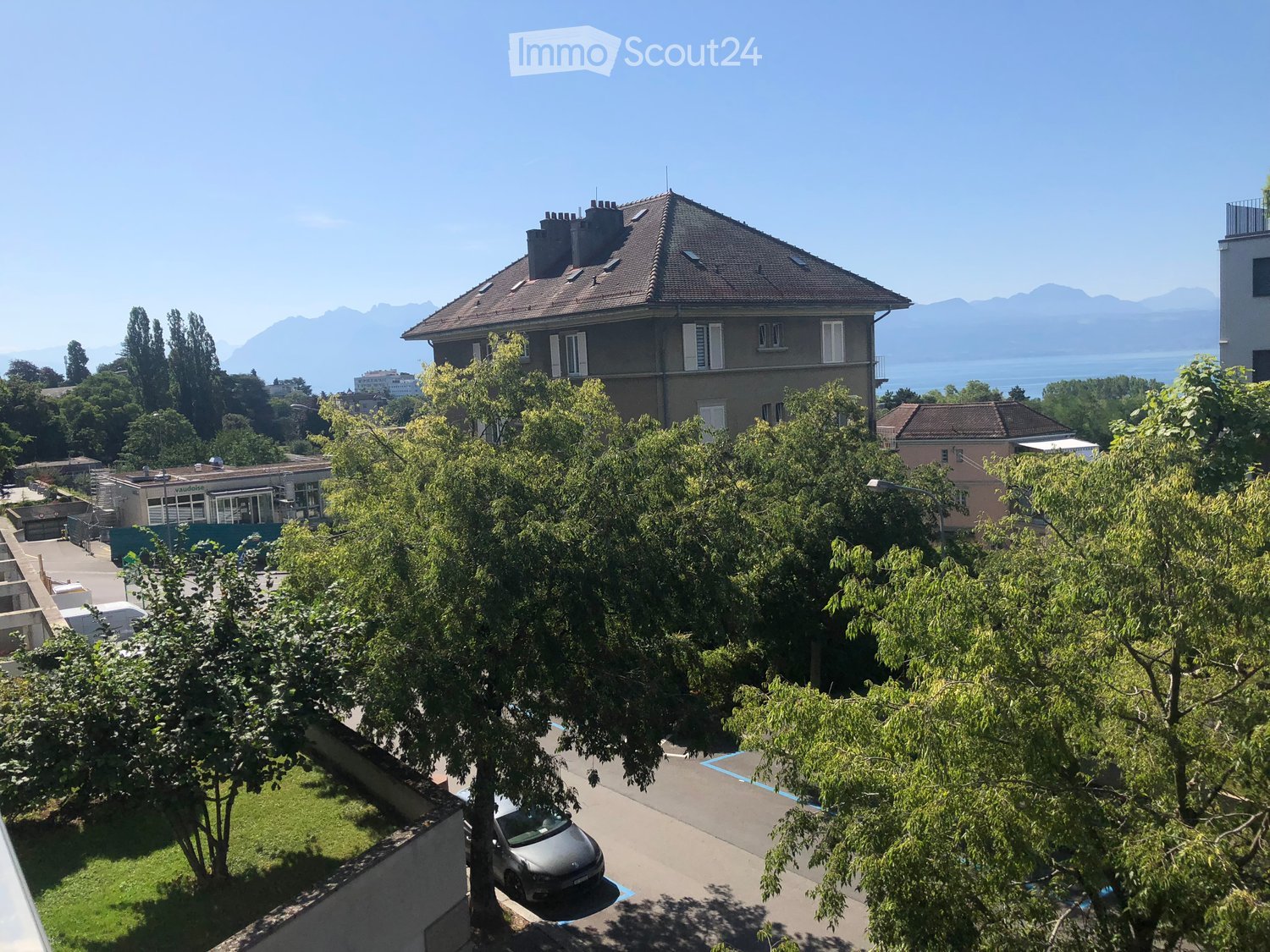 Balcony view of a residential area with trees, parked car, and distant mountains and lake.