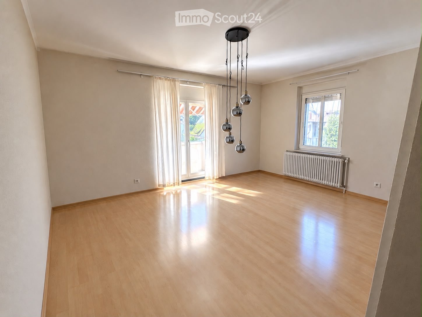 Empty room with wooden flooring, white walls, large windows with curtains, and a hanging light fixture.