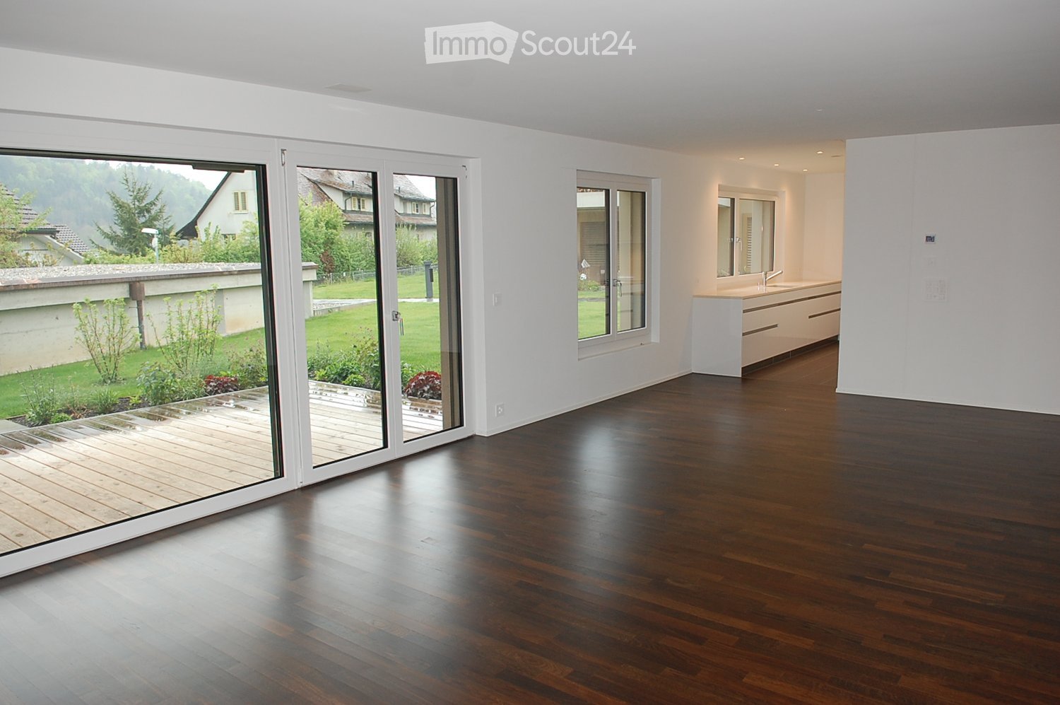 Spacious, unfurnished room with dark hardwood flooring, large sliding glass doors leading to a wooden deck, and a view of a green garden. A small, modern bathroom is visible in the background.