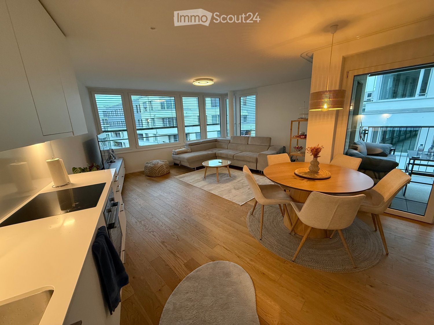 Open-plan living and dining area with modern furniture, large windows, and a balcony. Features a sofa, coffee table, ottoman, dining table with chairs, and a kitchen area with a counter and oven.