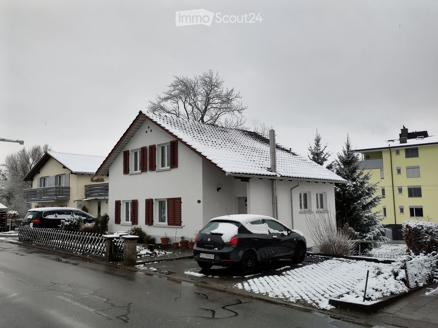 Two-story house with white exterior, red shutters, snow-covered roof, and a driveway with parked cars.