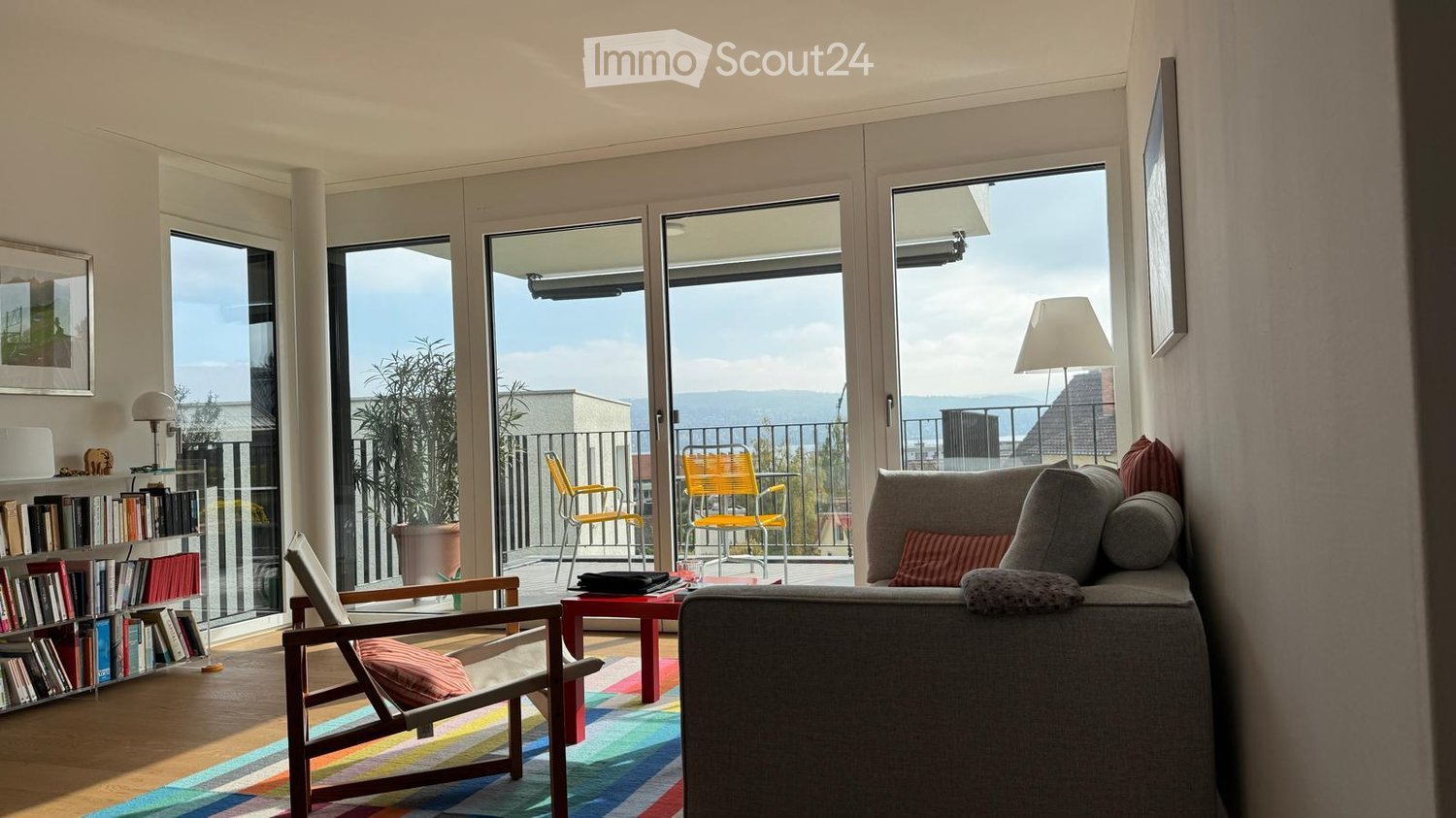 Bright living room with large windows and a balcony, featuring a grey sofa, wooden chair, red coffee table, bookshelf, and floor lamp.