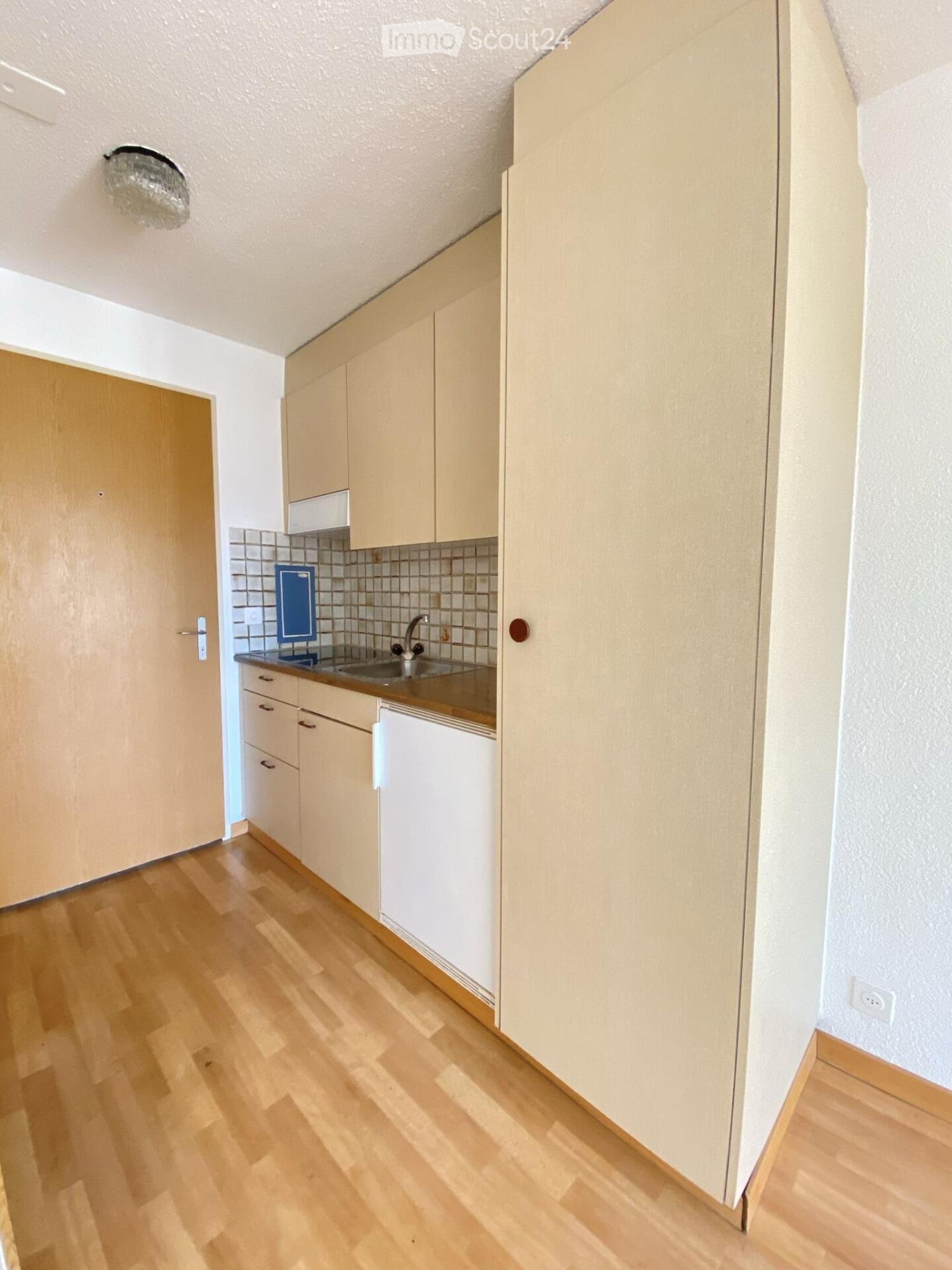 Modern kitchen with light wooden cabinets, a stainless steel sink, and a white refrigerator. The floor is made of light wooden planks.