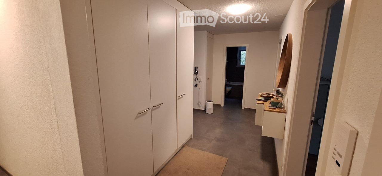 A narrow hallway with white walls, grey carpeted floor, and a large white wardrobe on the left. A small bathroom is visible at the end of the hallway, with a countertop, mirror, and various items on display.