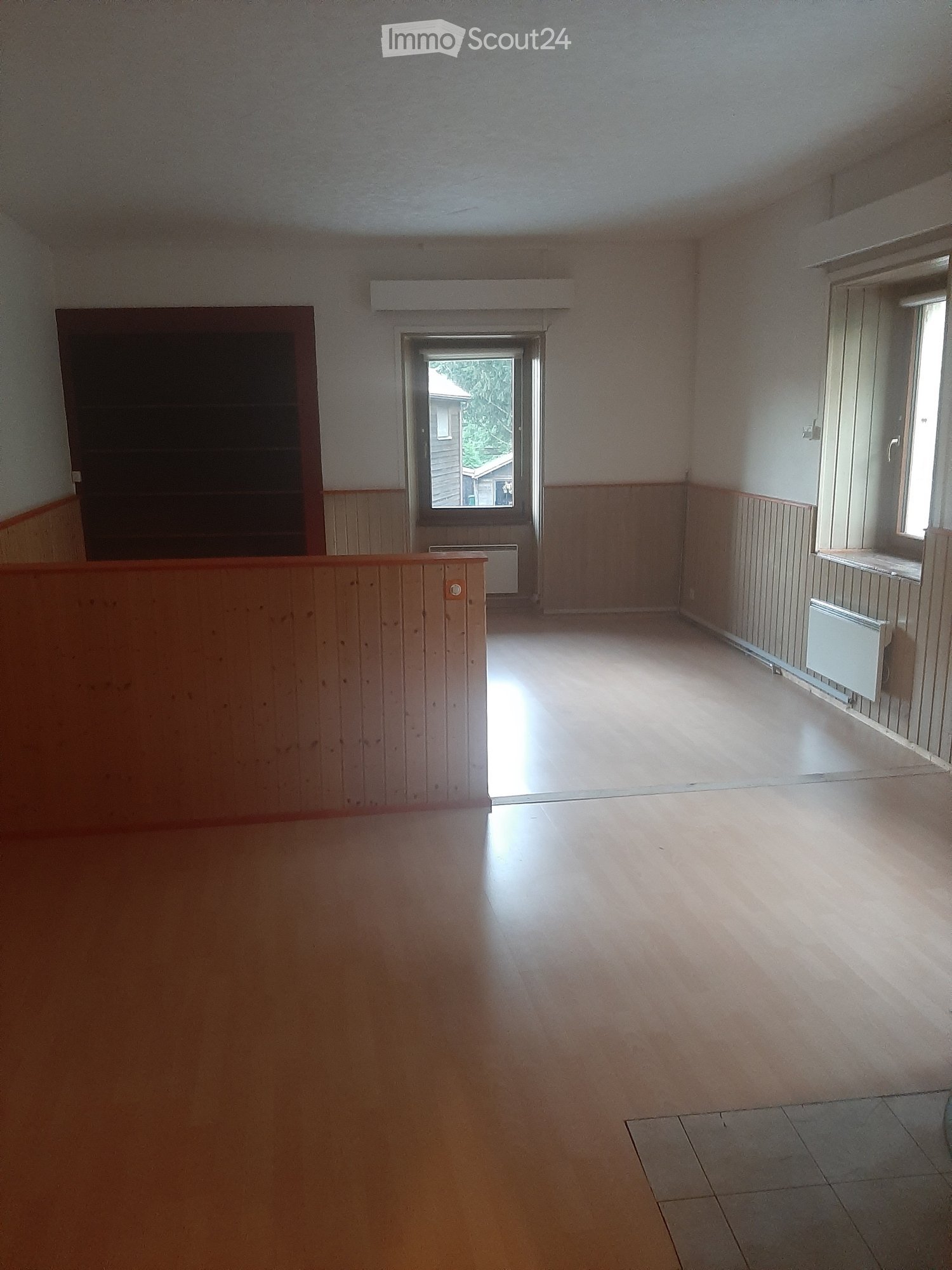 Empty room with wooden flooring, white walls, and a large window letting in natural light. There is a radiator on the wall and a doorway leading to another space.