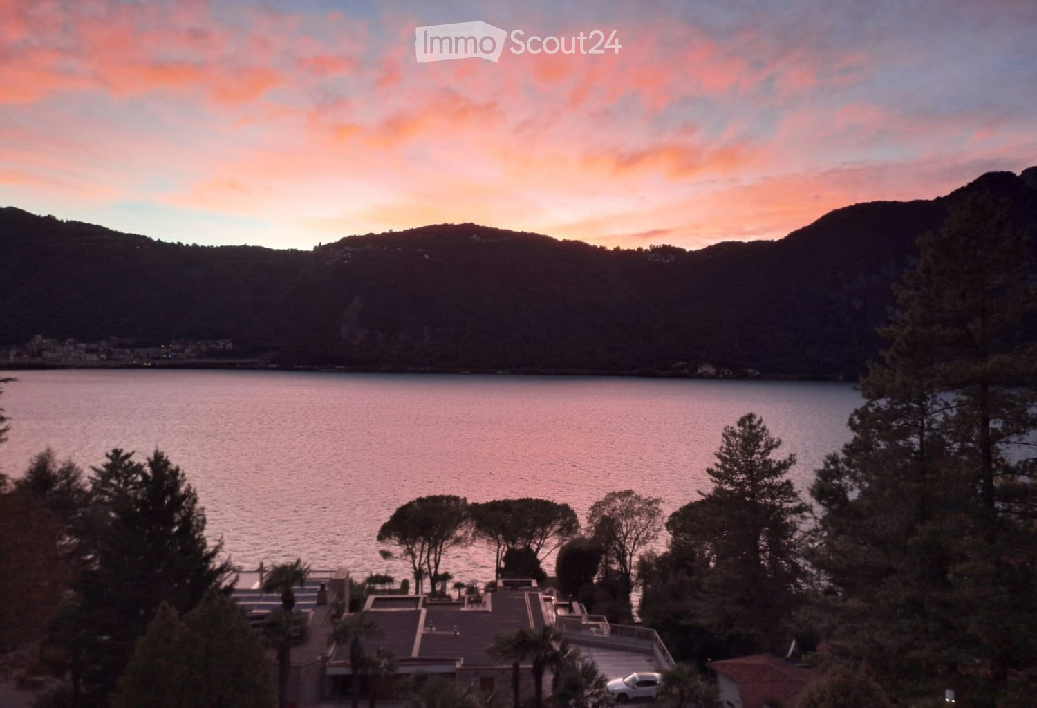 A scenic view of a lake surrounded by trees and mountains, with a house and parking area in the foreground.