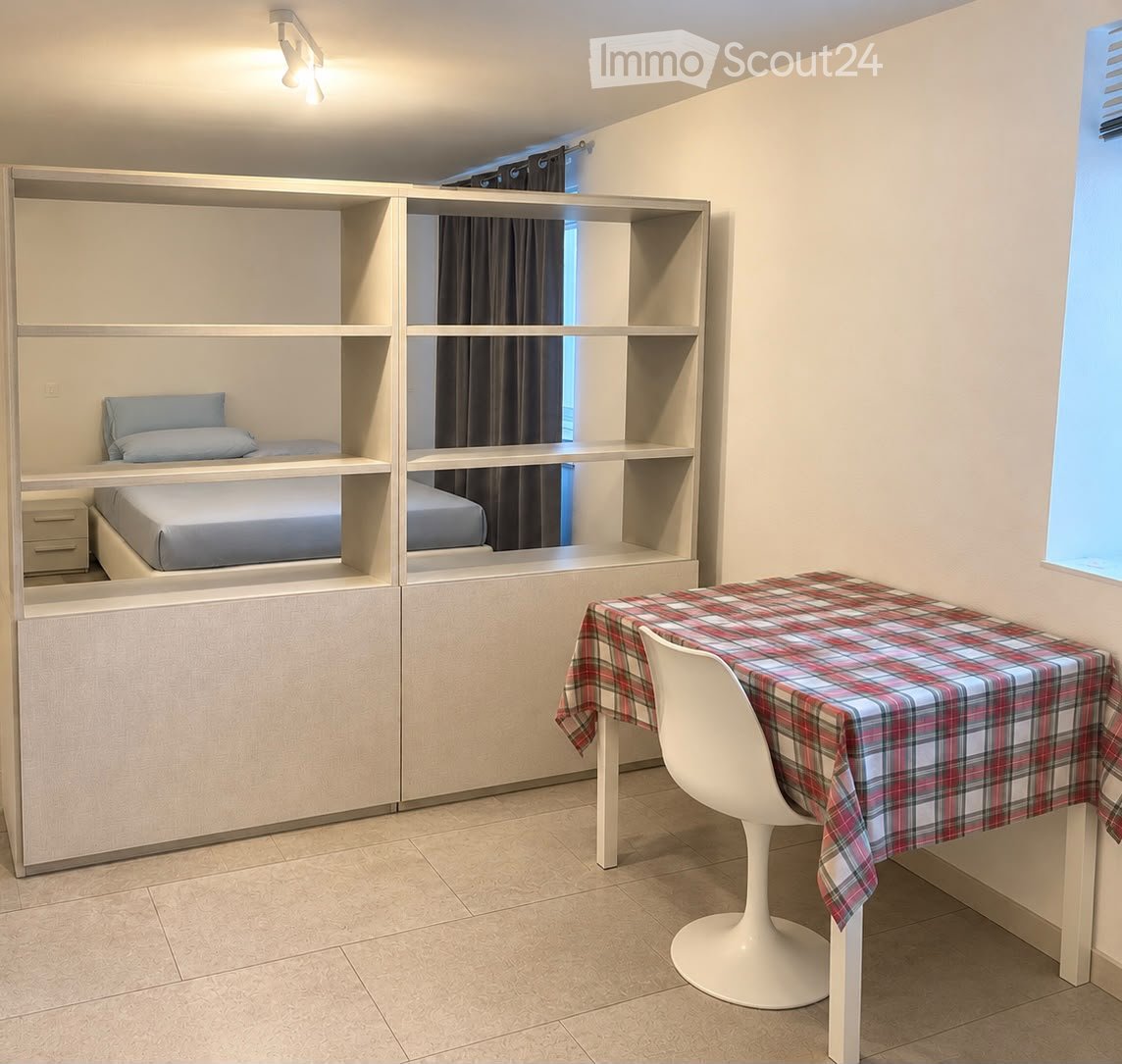A compact, modern bedroom with built-in bunk beds, a small table with a plaid tablecloth, and a white chair. The room features light-colored walls, tiled flooring, and built-in shelving units.