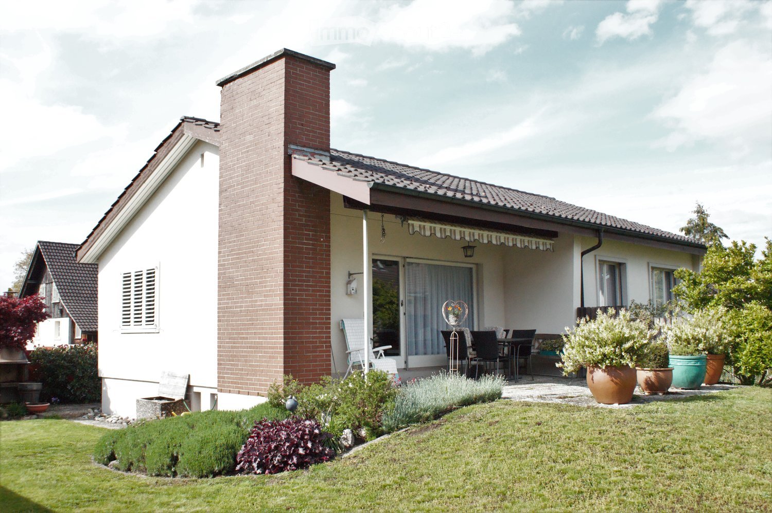 Two-story house with a brick chimney, white walls, and a covered patio area with outdoor furniture and potted plants.