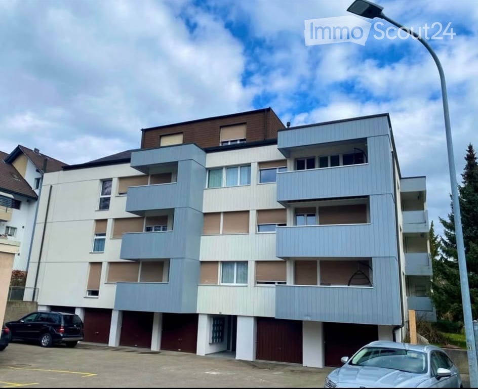 Modern multi-story apartment building with balconies, light blue and white facade, and parking area.