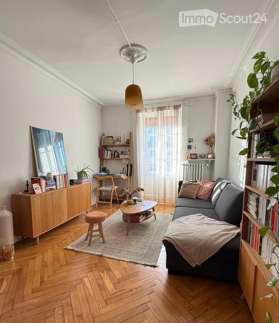 Modern living room with wooden herringbone flooring, a grey sectional sofa, a round coffee table, a small wooden stool, built-in bookshelves, a wooden cabinet, and decorative plants.