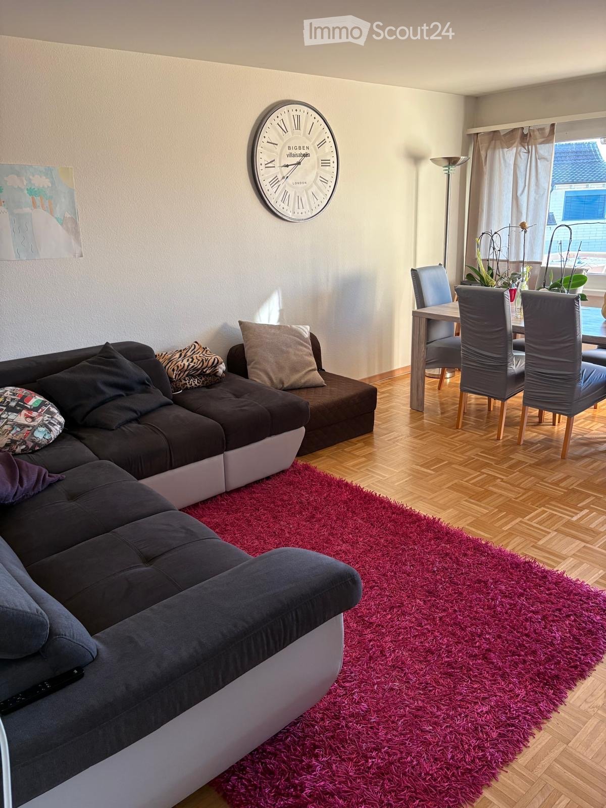 Modern living room with a dark grey sectional sofa, colorful throw pillows, a vibrant red shag rug, a dining area with chairs and a table, a large wall clock, and a floor lamp near the window.