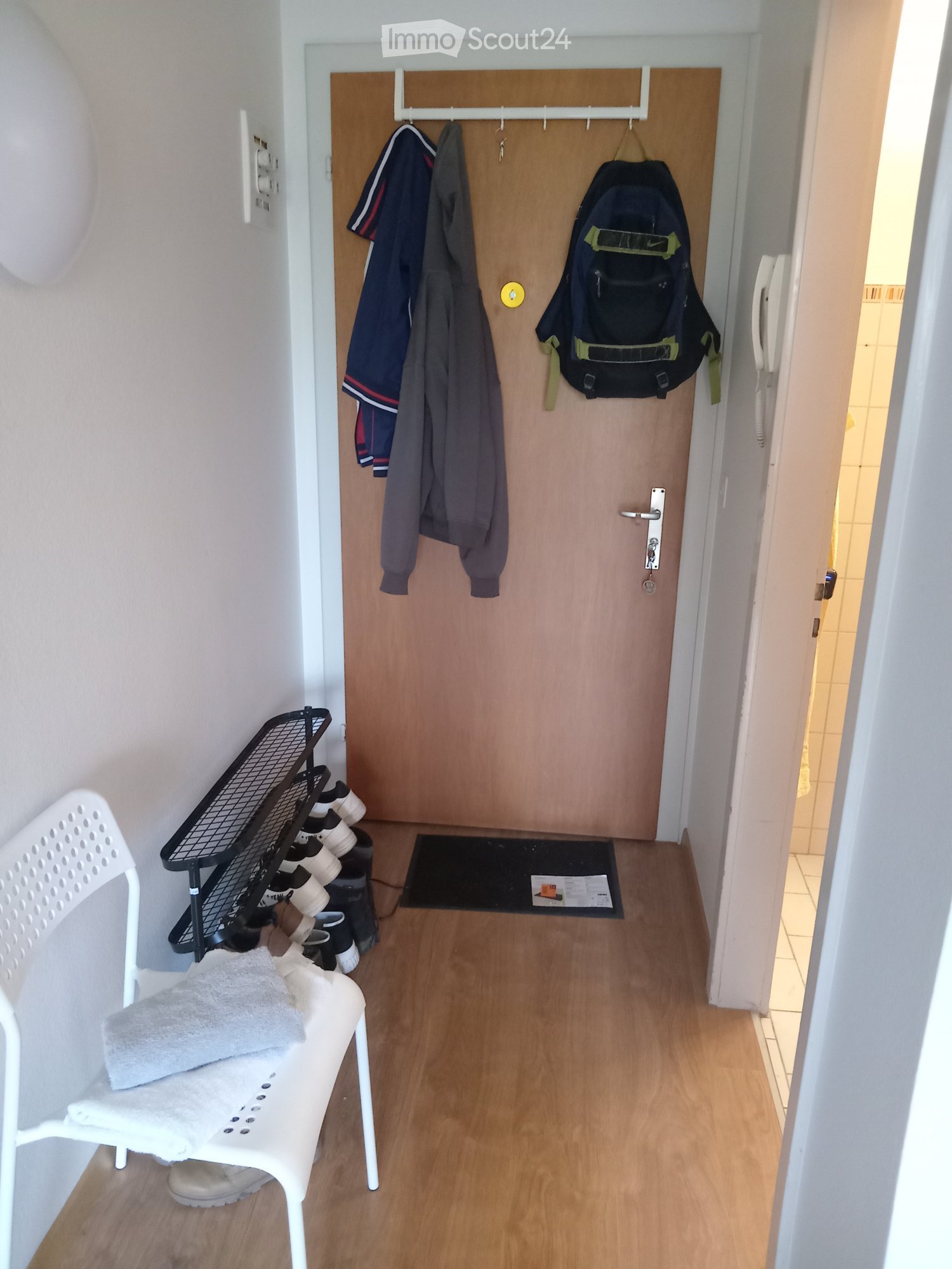 A narrow hallway with wooden flooring, a white plastic chair with a towel, a shoe rack with several pairs of shoes, a coat rack with jackets and a backpack, and a small mat in front of a wooden door.