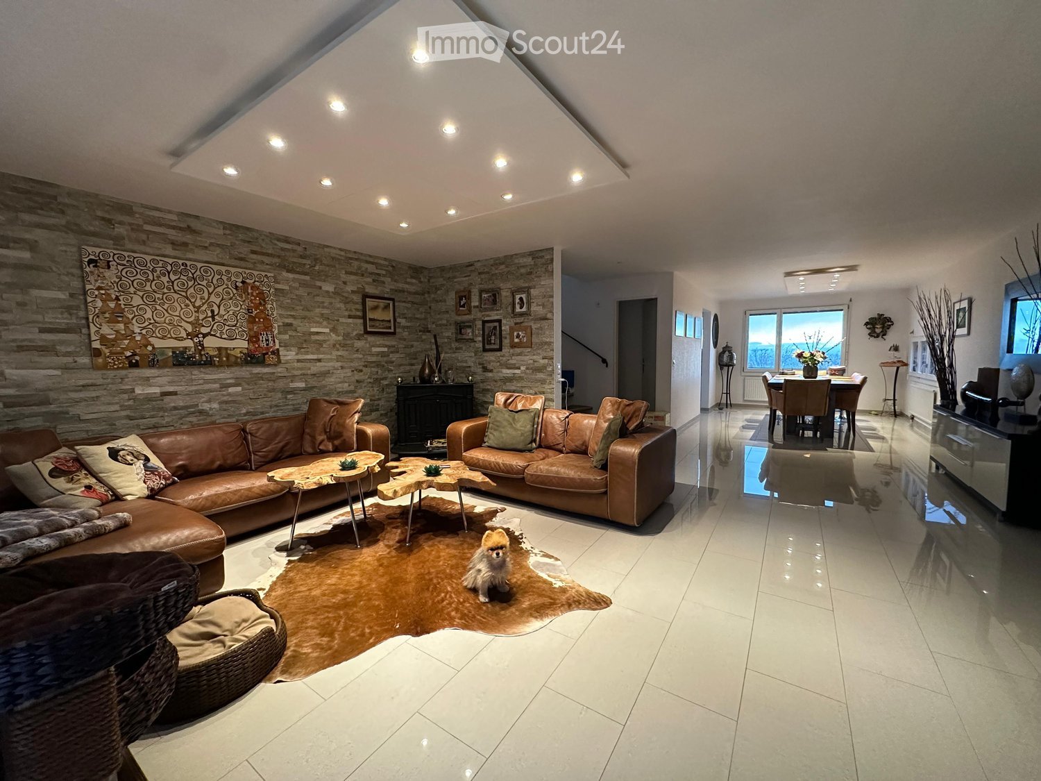Modern living room with leather sofas, coffee tables, a fur rug, and a small dog. The room features a stone accent wall, recessed lighting, and a view of a dining area through an open doorway.