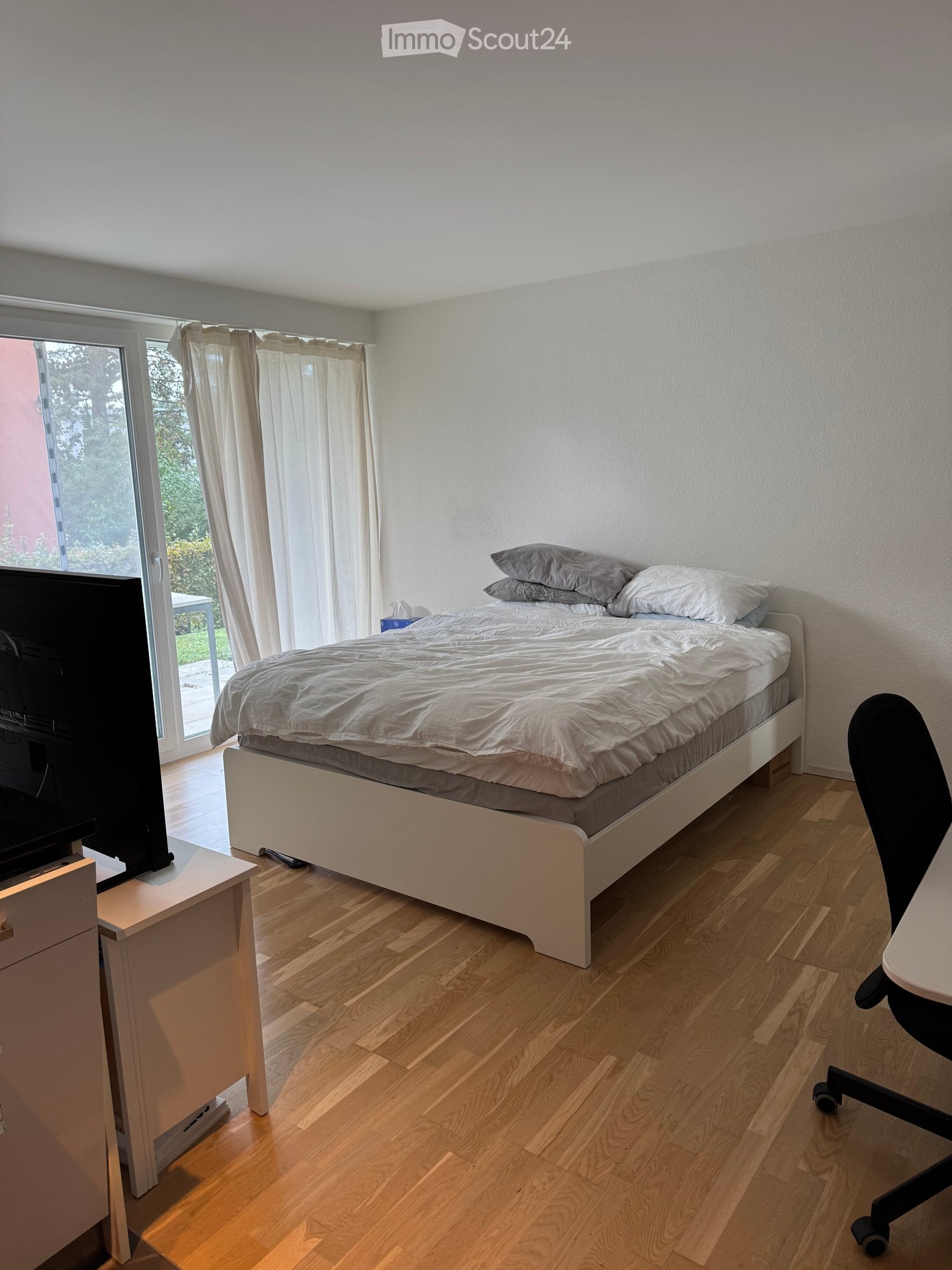 A modern bedroom with a large bed, pillows, a TV on a stand, a chair, and a desk with a chair.