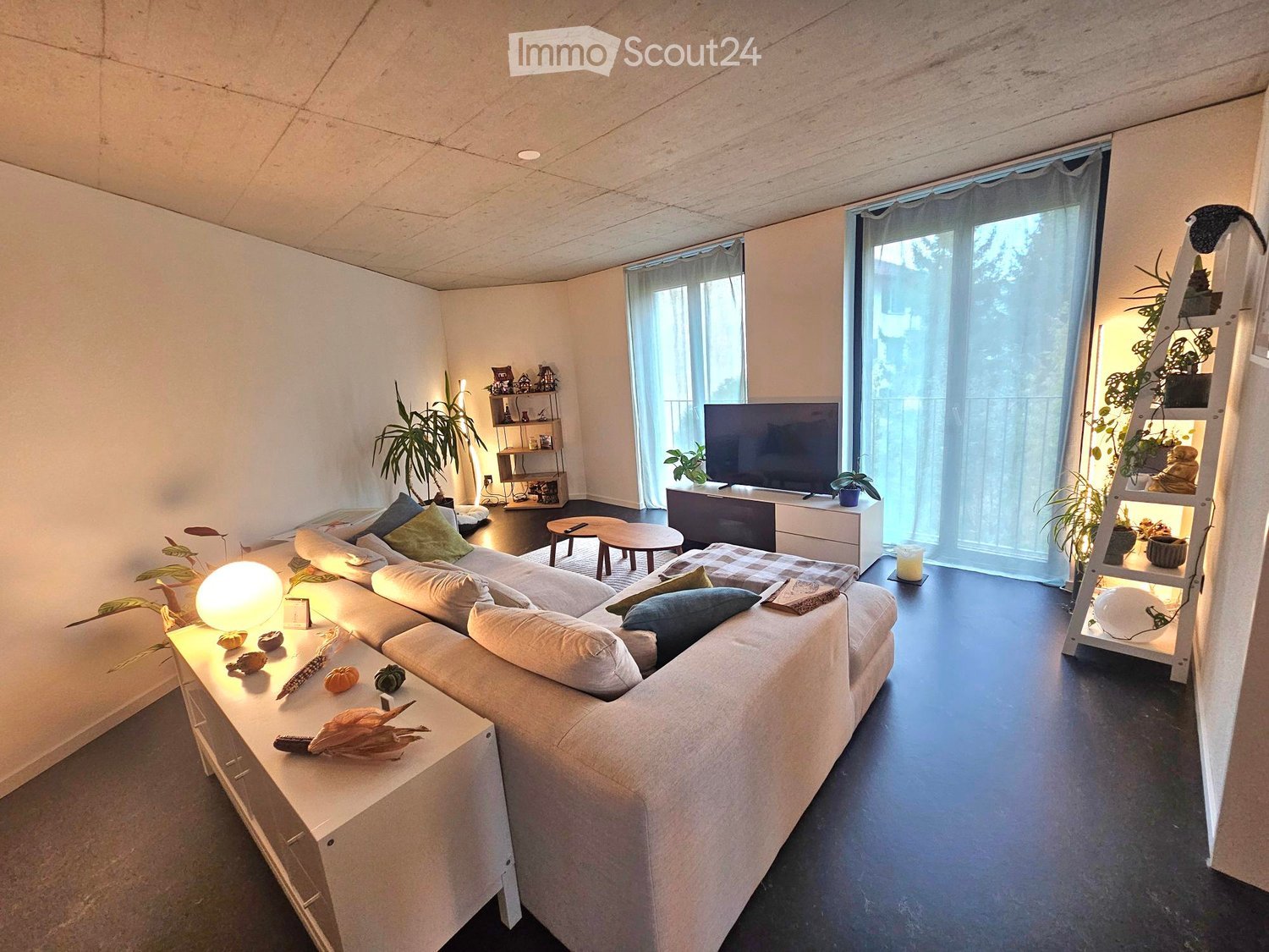 Modern living room with a large sectional sofa, coffee table, side tables, TV, shelves, and decorative plants.