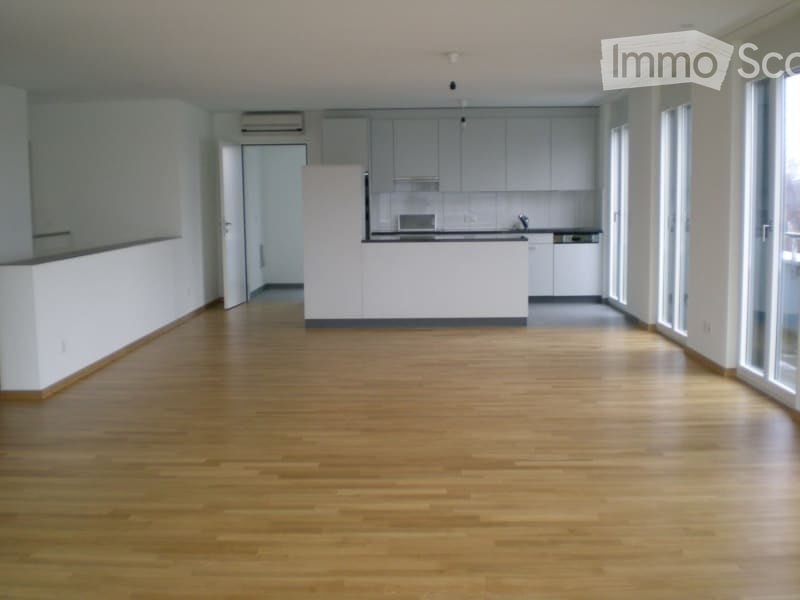 Empty room with wooden flooring, white walls, and a white kitchen area in the background. No furniture or equipment is present.