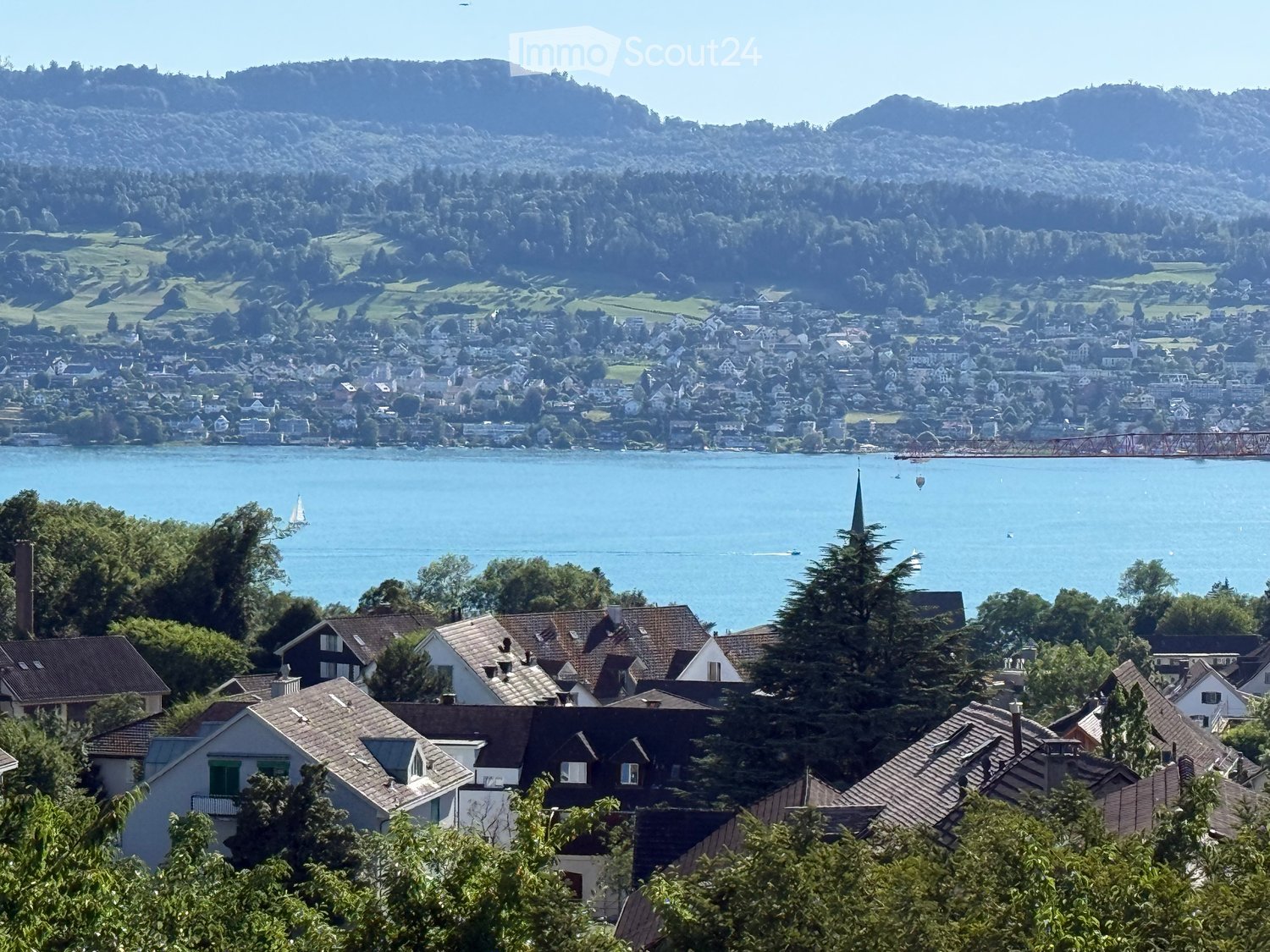 A scenic view of a town with houses, a lake, and surrounding hills. The town features numerous residential buildings with varied roof styles, surrounded by lush greenery and a calm lake with sailboats. In the background, there are rolling hills and a dist