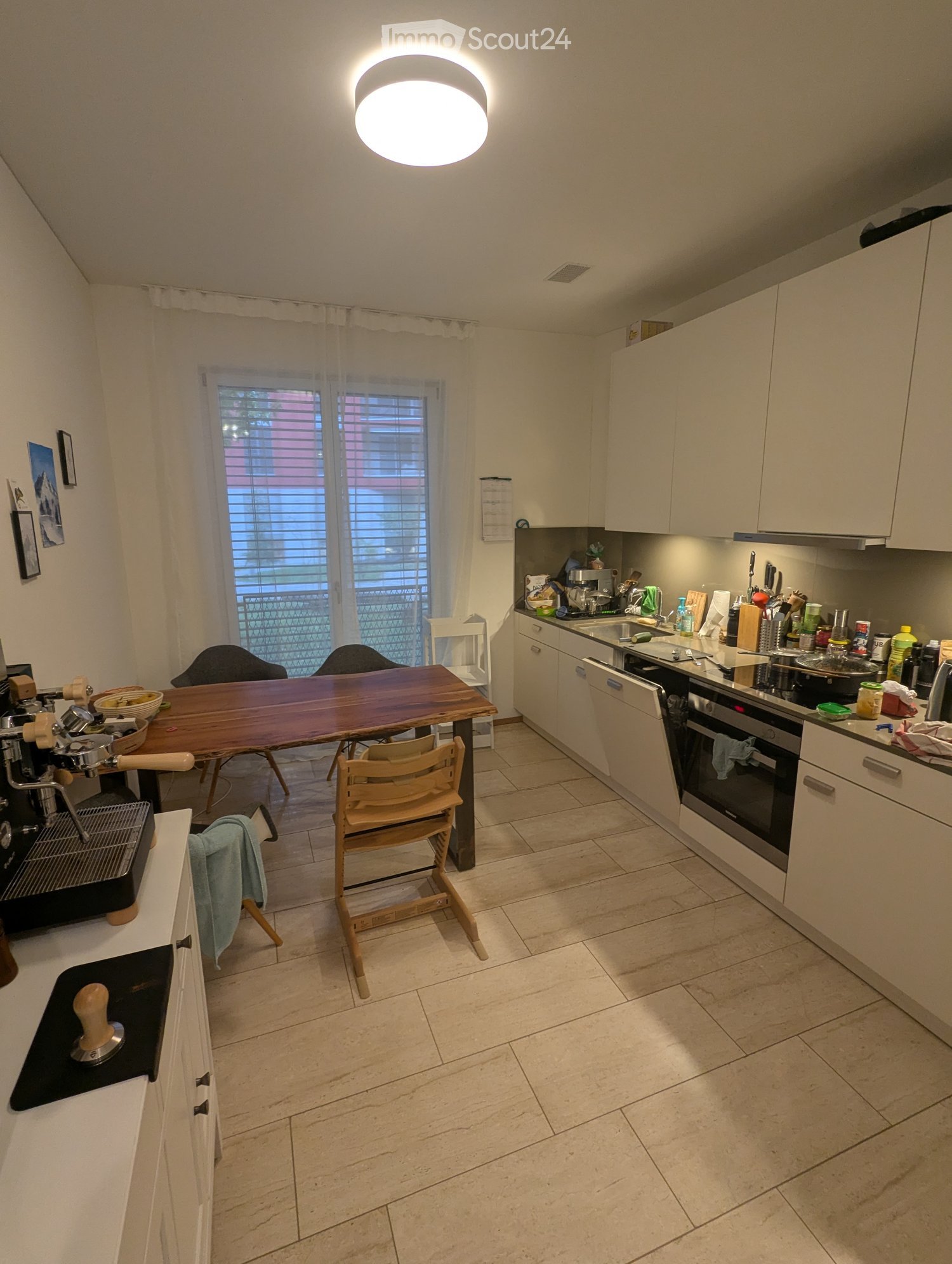 Modern kitchen with white cabinets, a wooden dining table, and various appliances including an oven, dishwasher, and coffee machine.