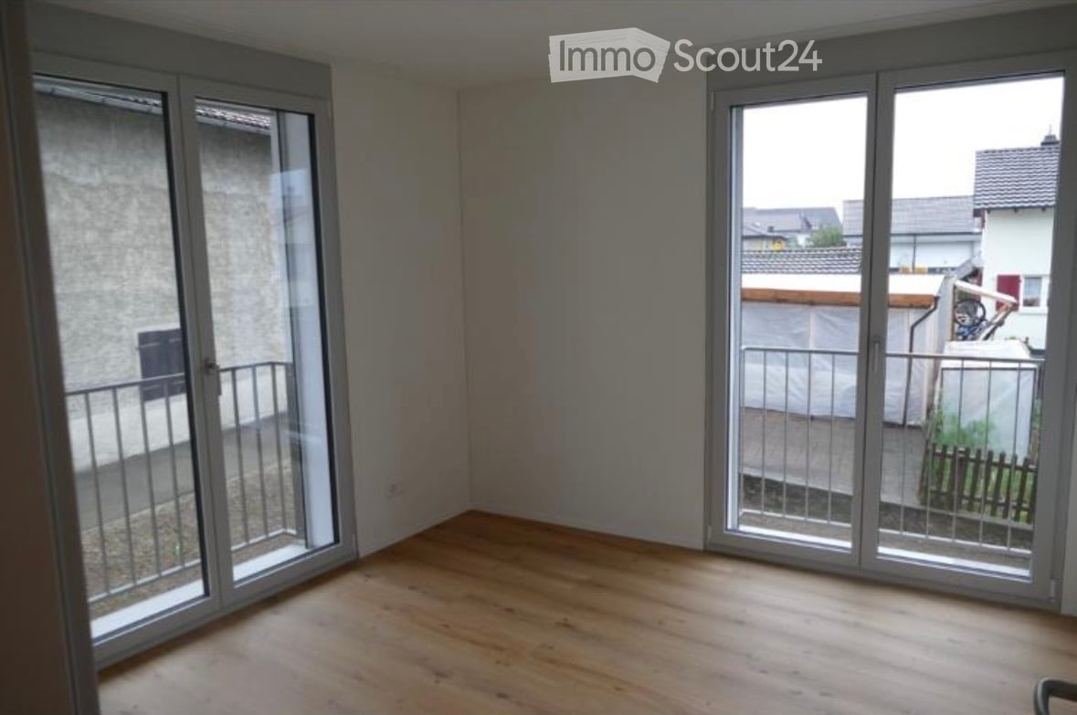 Empty room with wooden flooring, large sliding glass doors leading to a balcony, and a view of neighboring buildings.