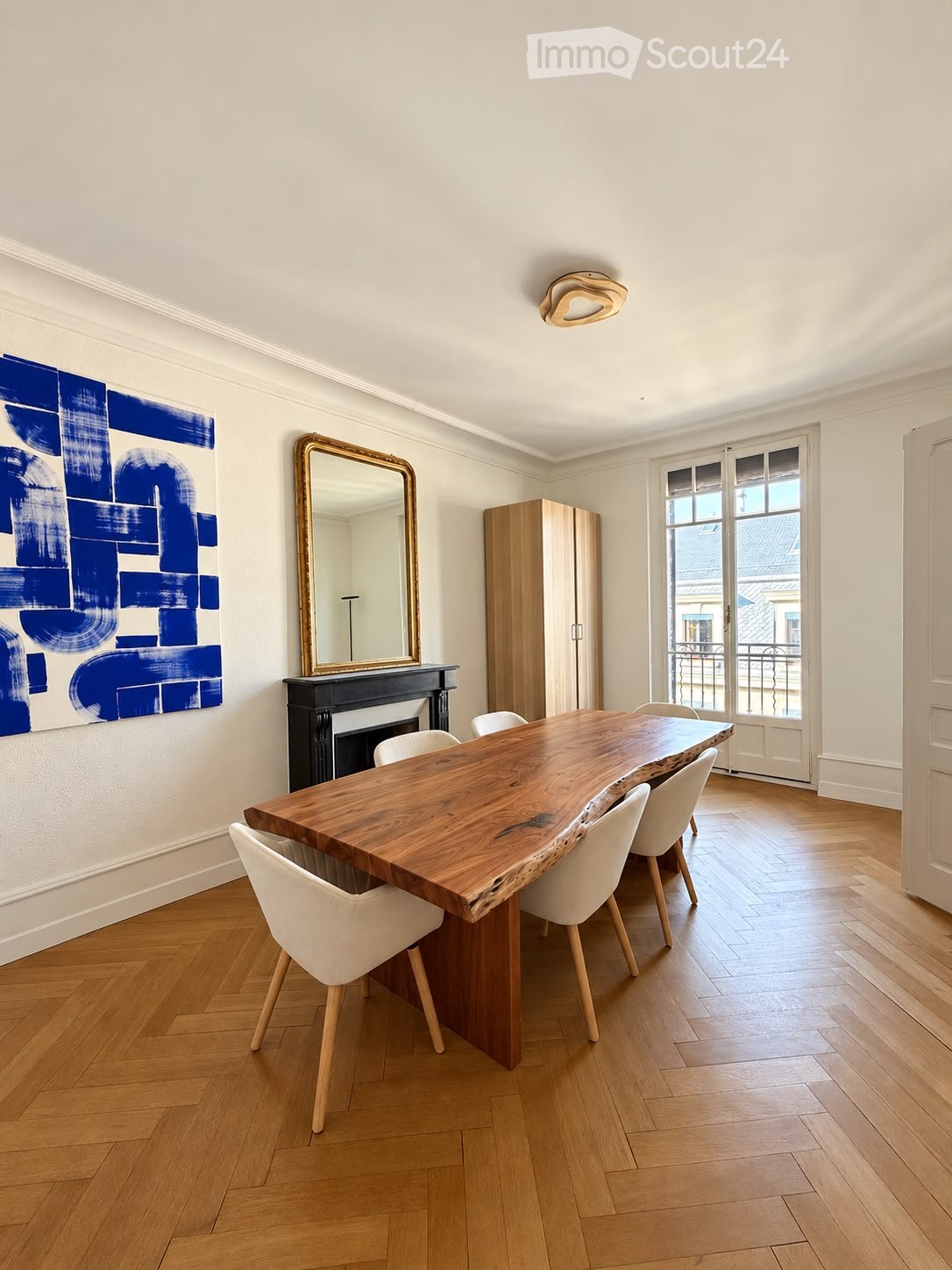 Dining room with a large wooden dining table, white chairs, a mirror, a cabinet, and a fireplace with artwork on the wall.