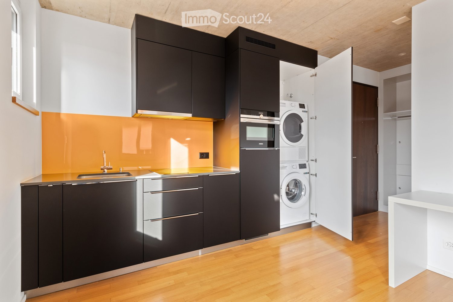 Modern kitchen with dark cabinets, stainless steel appliances, and a bright orange backsplash. Features a sink, oven, and washing machine.