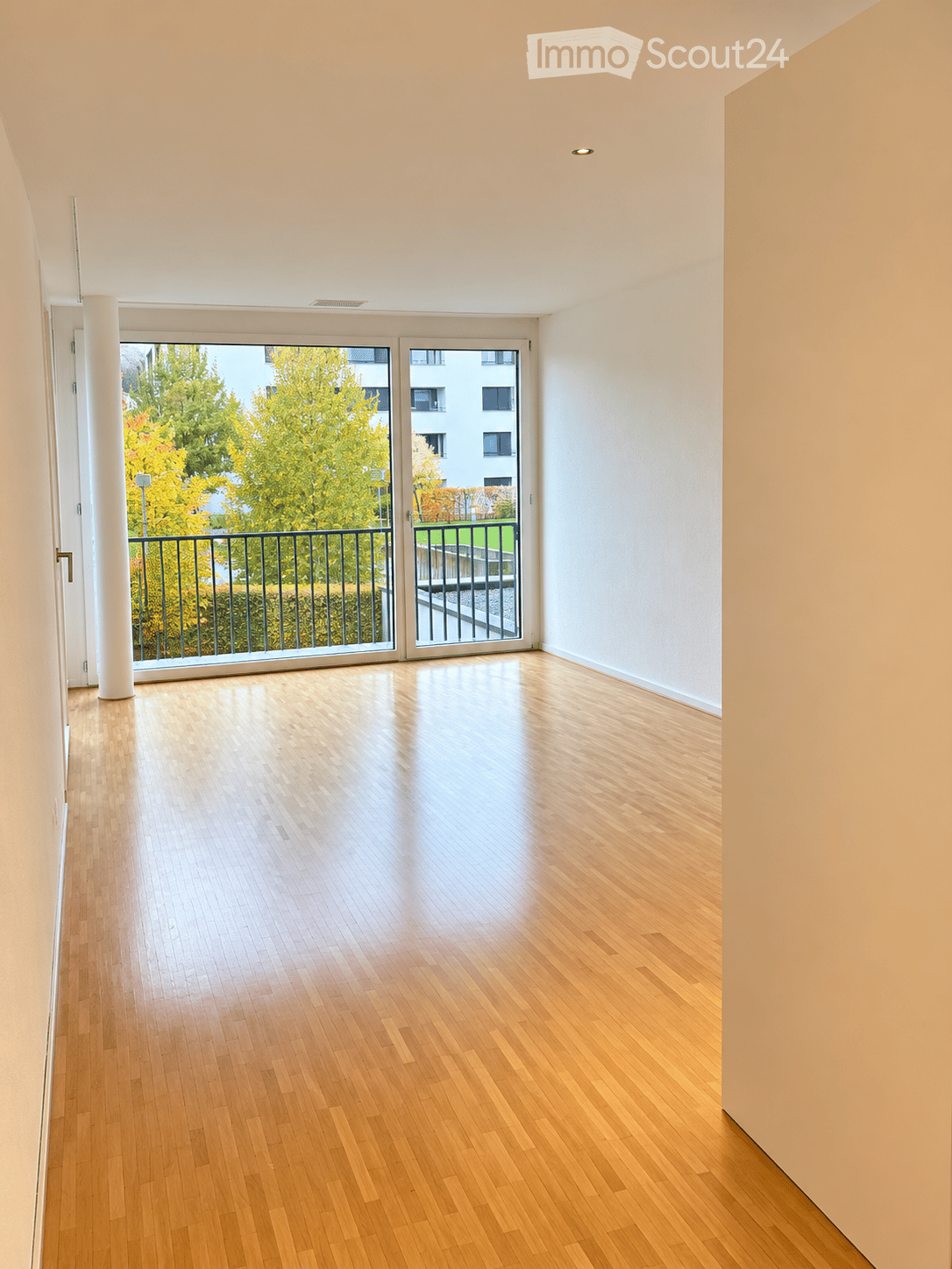 Empty room with light wooden flooring, white walls, and large sliding glass doors leading to a balcony with a metal railing and view of greenery.