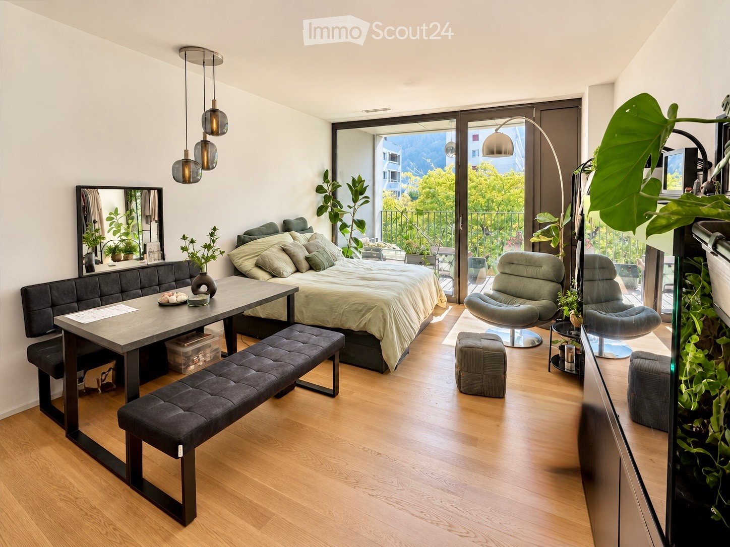 Modern, open-concept living and sleeping area with a bed, sofa, dining table, chairs, and several stools. Large windows and sliding glass doors lead to a balcony with outdoor seating.