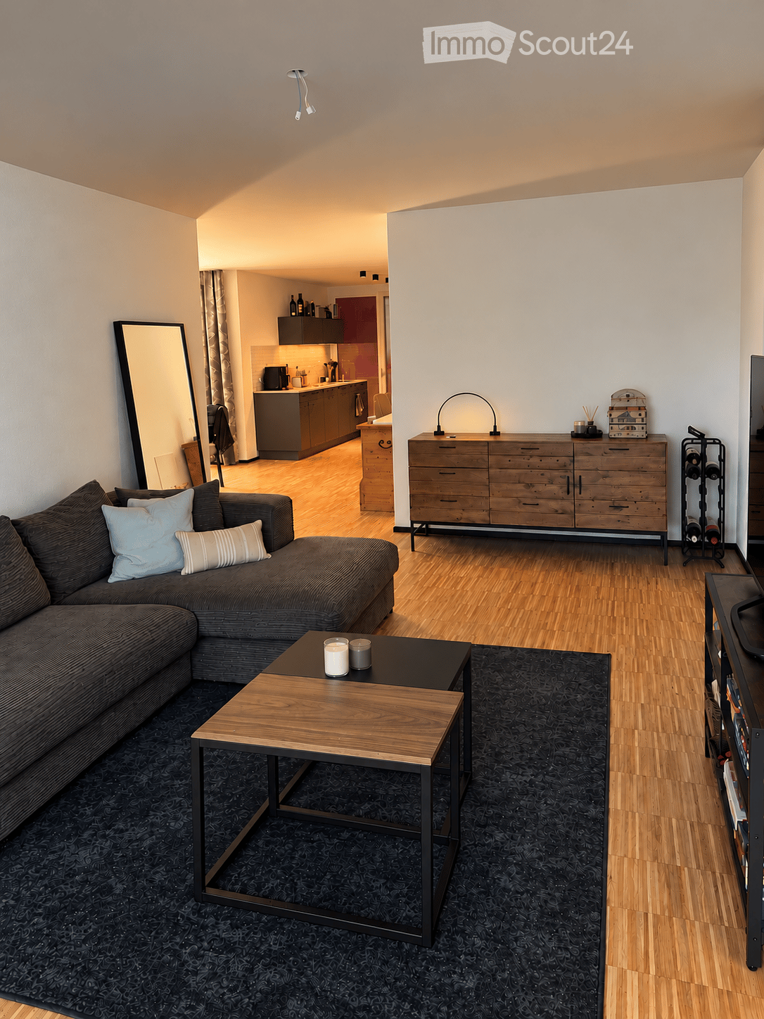 Modern living room with a sectional sofa, coffee table, and a large mirror. The room features a wooden dresser, a wine rack, and a television. An open kitchen area is visible in the background with cabinets, a counter, and various appliances.