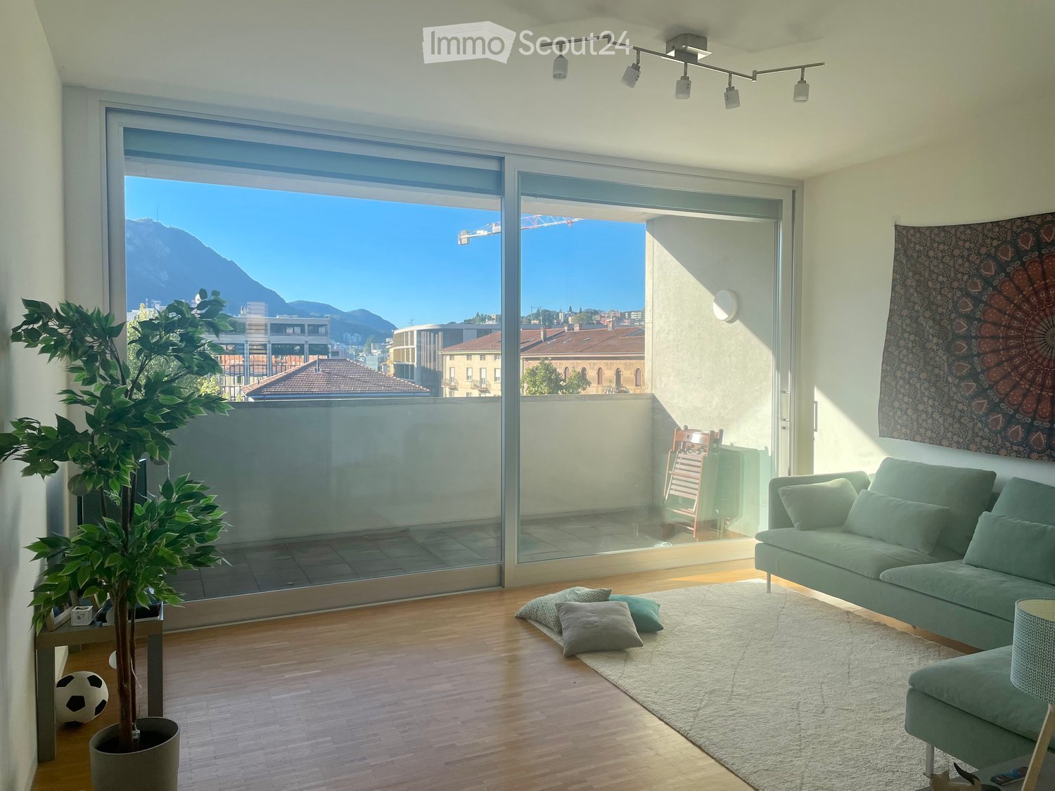 Modern living room with large glass sliding doors leading to a balcony, offering a scenic mountain view. Features a green sofa, throw pillows, a potted plant, and a decorative wall hanging.