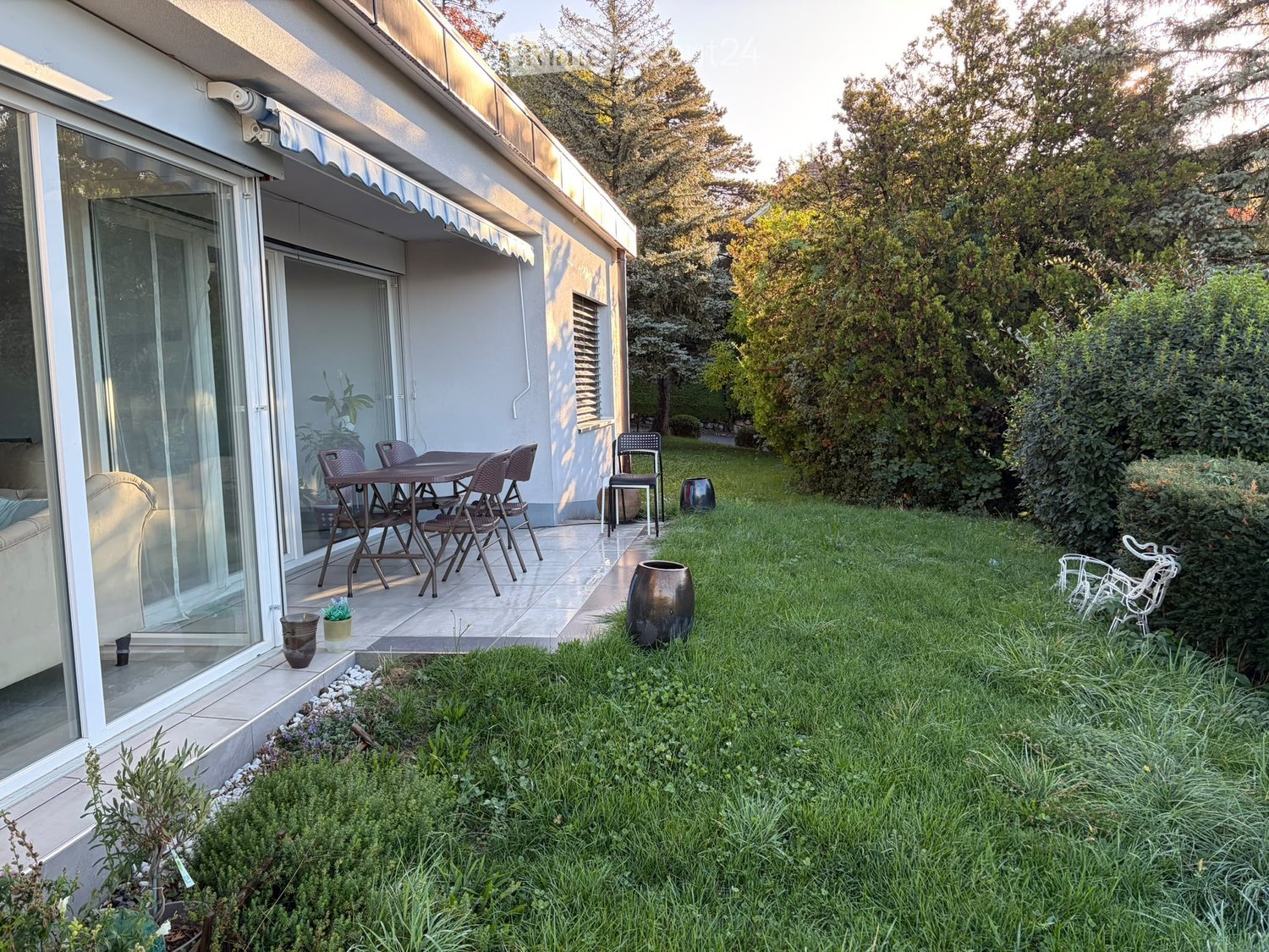 Garden with a patio area featuring a table and chairs, surrounded by greenery and decorative vases.