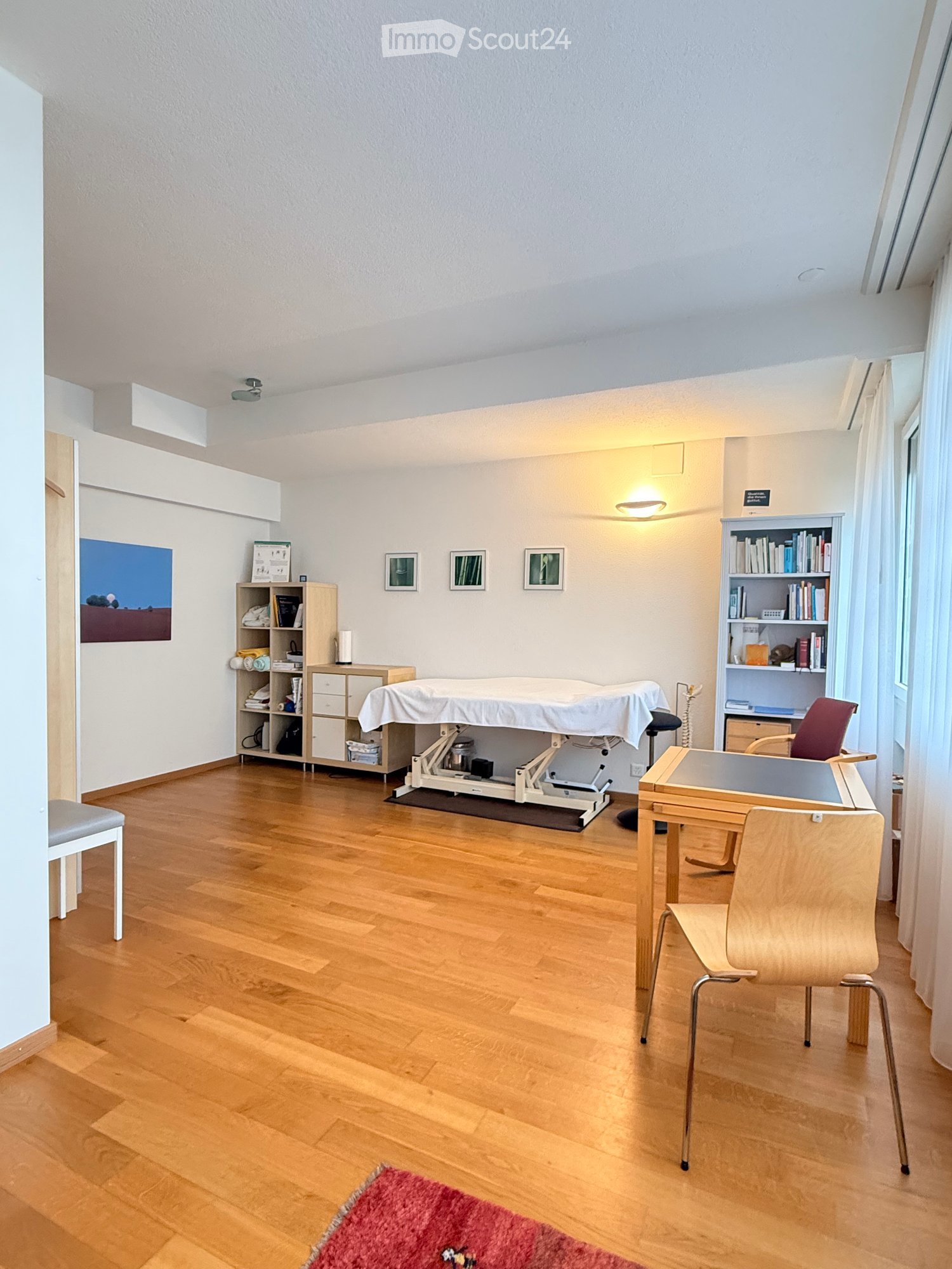 A well-lit room with wooden flooring, a massage table, a small table with chairs, shelves, and a cabinet.