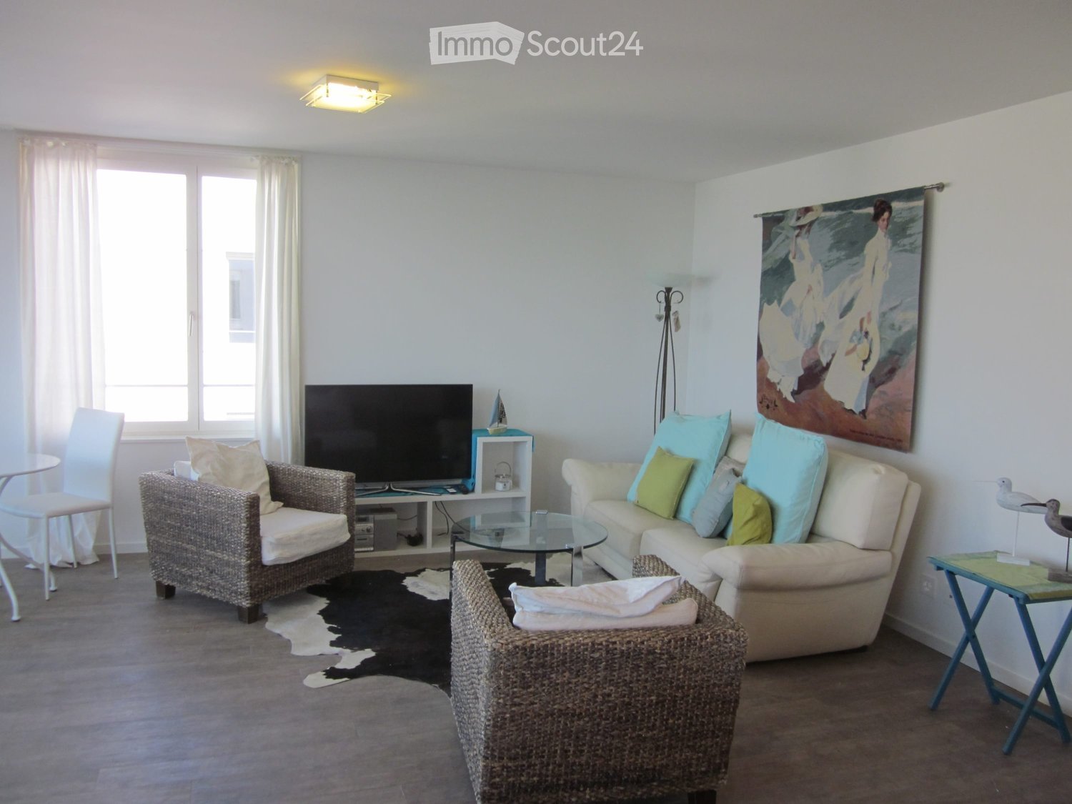 Bright living room with a cream-colored sofa, wicker chairs, a glass coffee table, a television, and a painting on the wall. The room features large windows with white curtains and a cowhide rug on the floor.