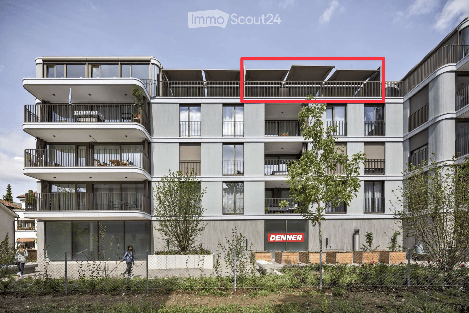 Modern multi-story residential building with balconies, large windows, and contemporary design. Features include a rooftop terrace with outdoor seating and a fenced garden area at the base.