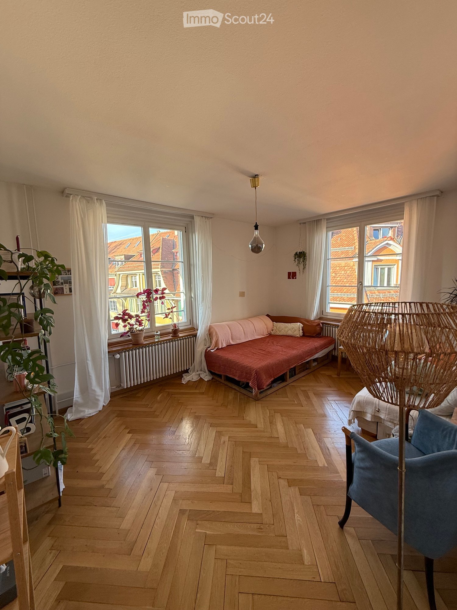 A cozy living room with wooden herringbone flooring, large windows with white curtains, a red sofa, a blue armchair, a woven floor lamp, and several potted plants.