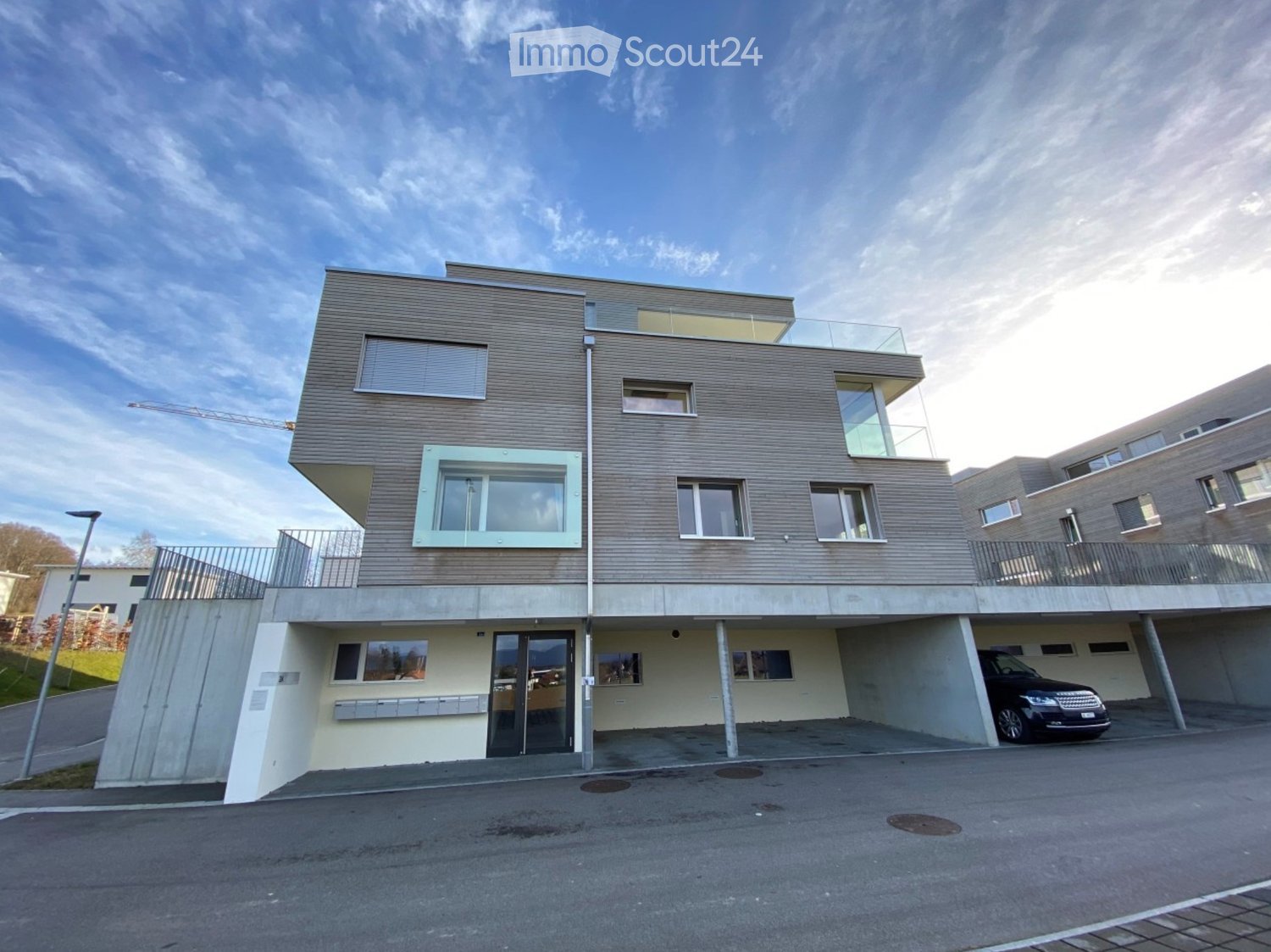 Modern two-story building with a raised ground floor, large windows, and a balcony on the upper level. A car is parked in the covered garage area.