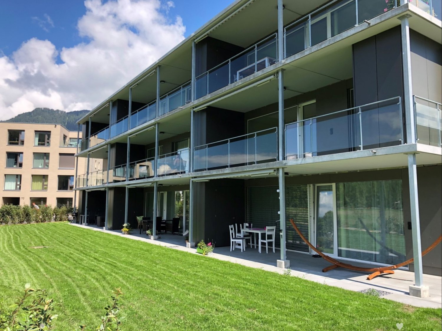 Modern multi-story building with balconies and a well-maintained lawn in front. Outdoor seating area with a table, chairs, and a sun lounger.
